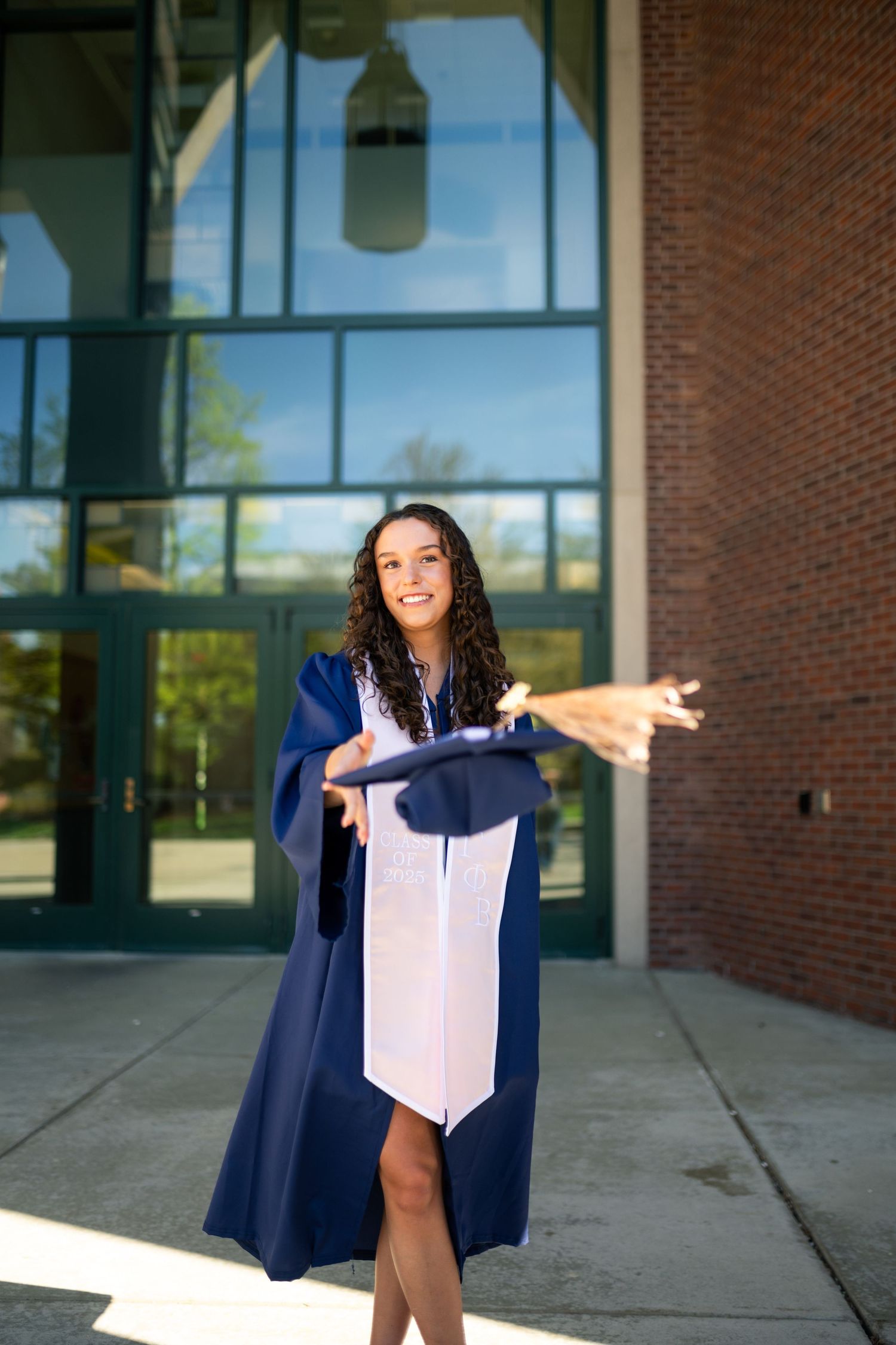 Graduate in navy blue cap and gown strikes a joyful pose outside brick building on sunny graduation day.