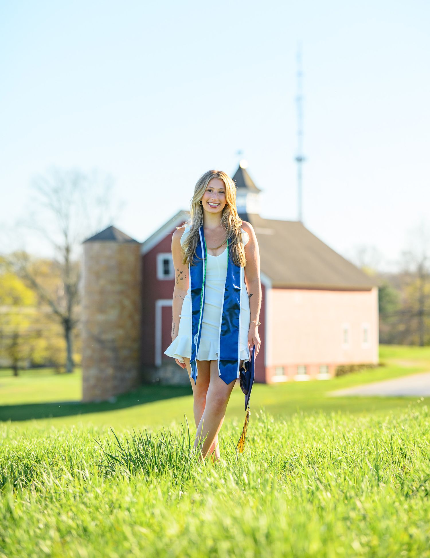 Someone in a blue dress stands in tall grass with a rustic red barn and silo in the background on a sunny day.