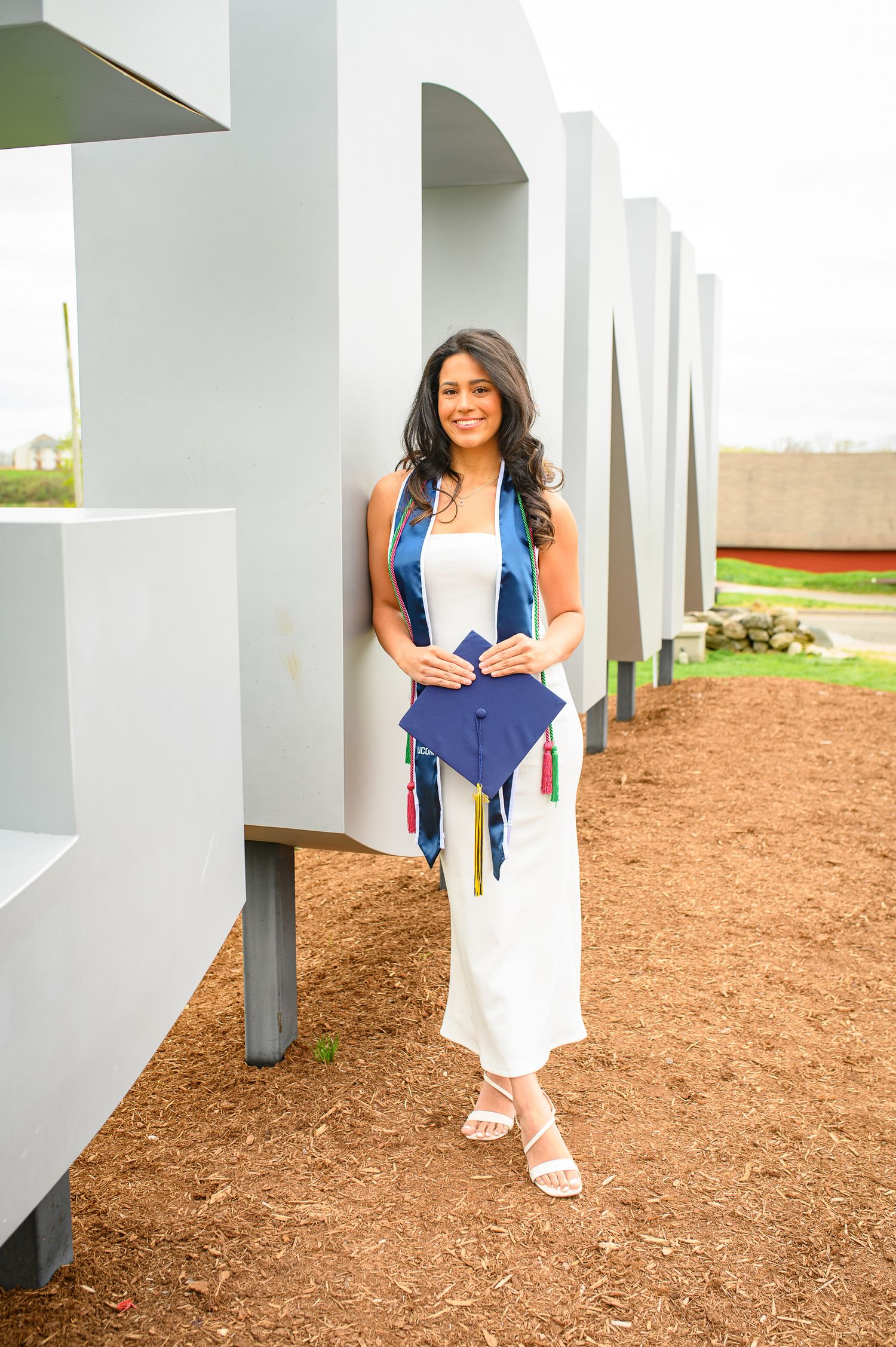 Graduate posing in white dress with blue academic stole standing between white architectural columns outdoors.