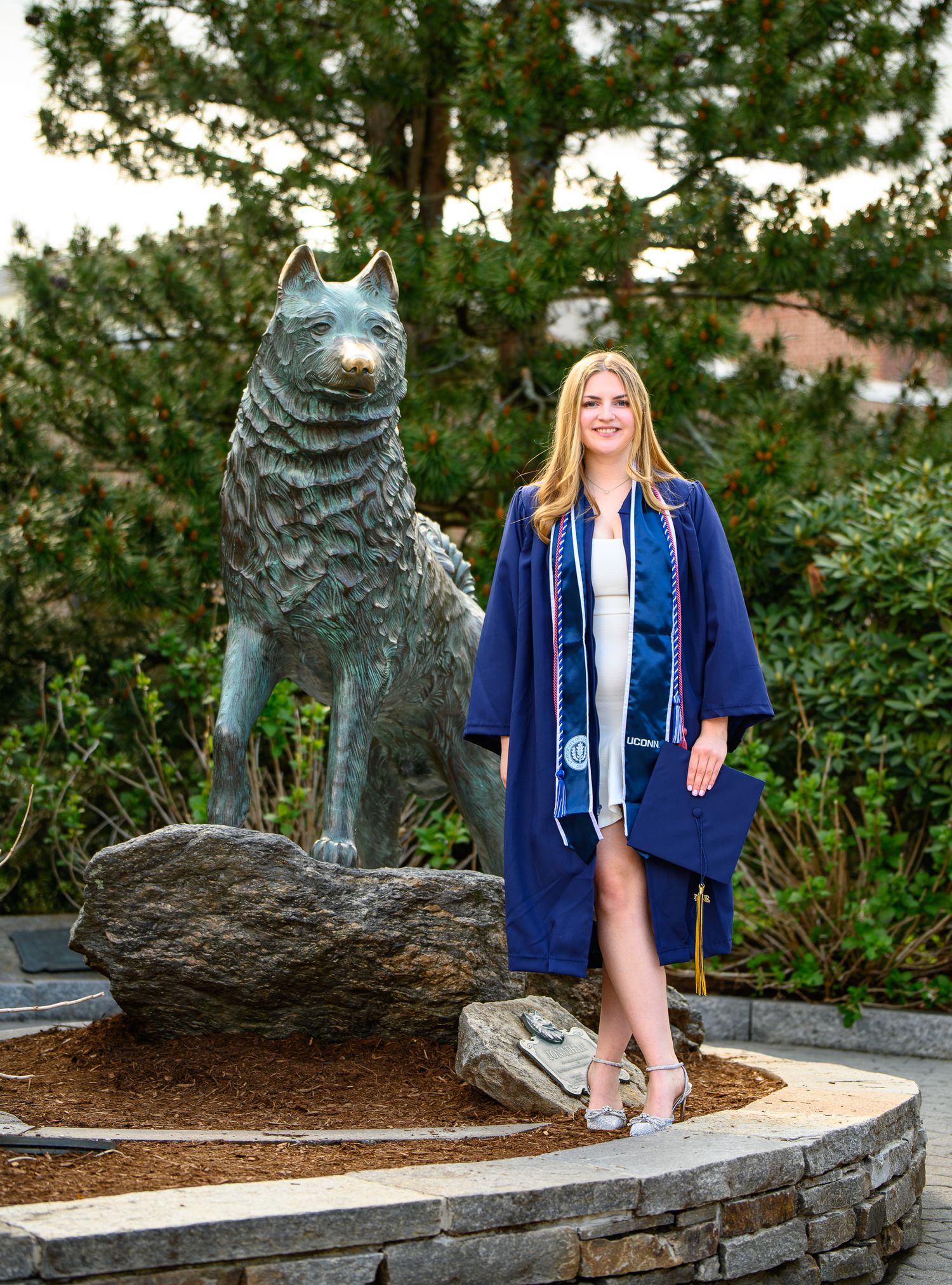 Graduate wearing blue commencement gown poses next to bronze husky mascot statue on stone platform outdoors.