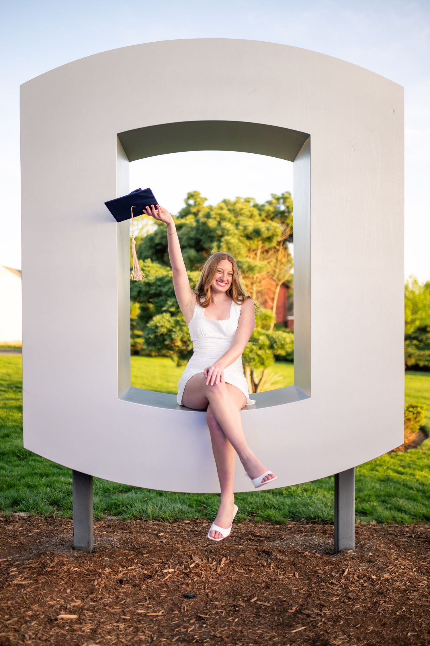 Graduate in white dress celebrates with diploma while sitting in decorative white architectural frame outdoors at sunset.