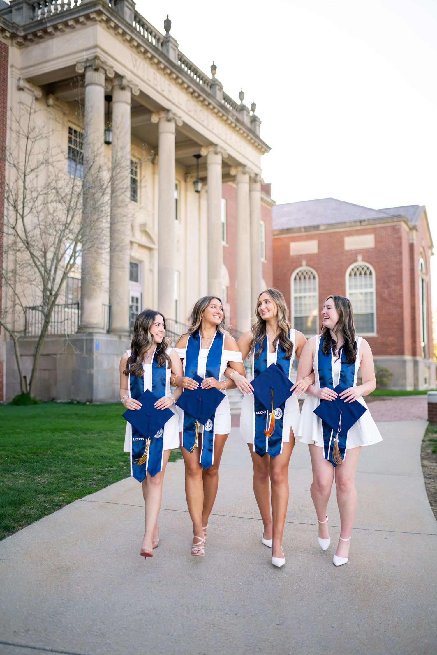 Four graduates in blue caps and gowns walking together along campus path in front of neoclassical university building.