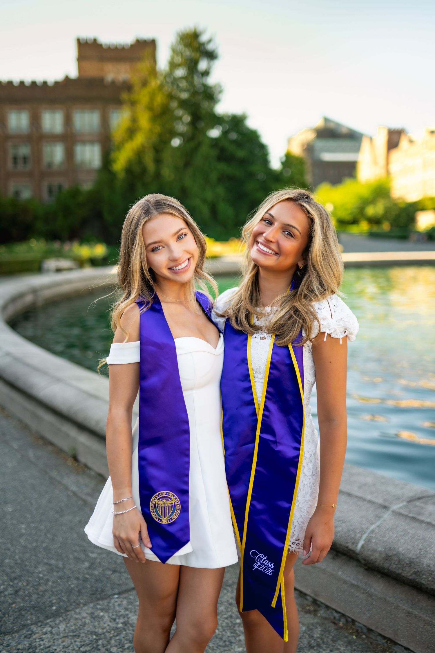 Two smiling graduates wearing purple stoles pose by a fountain on a sunny college campus.