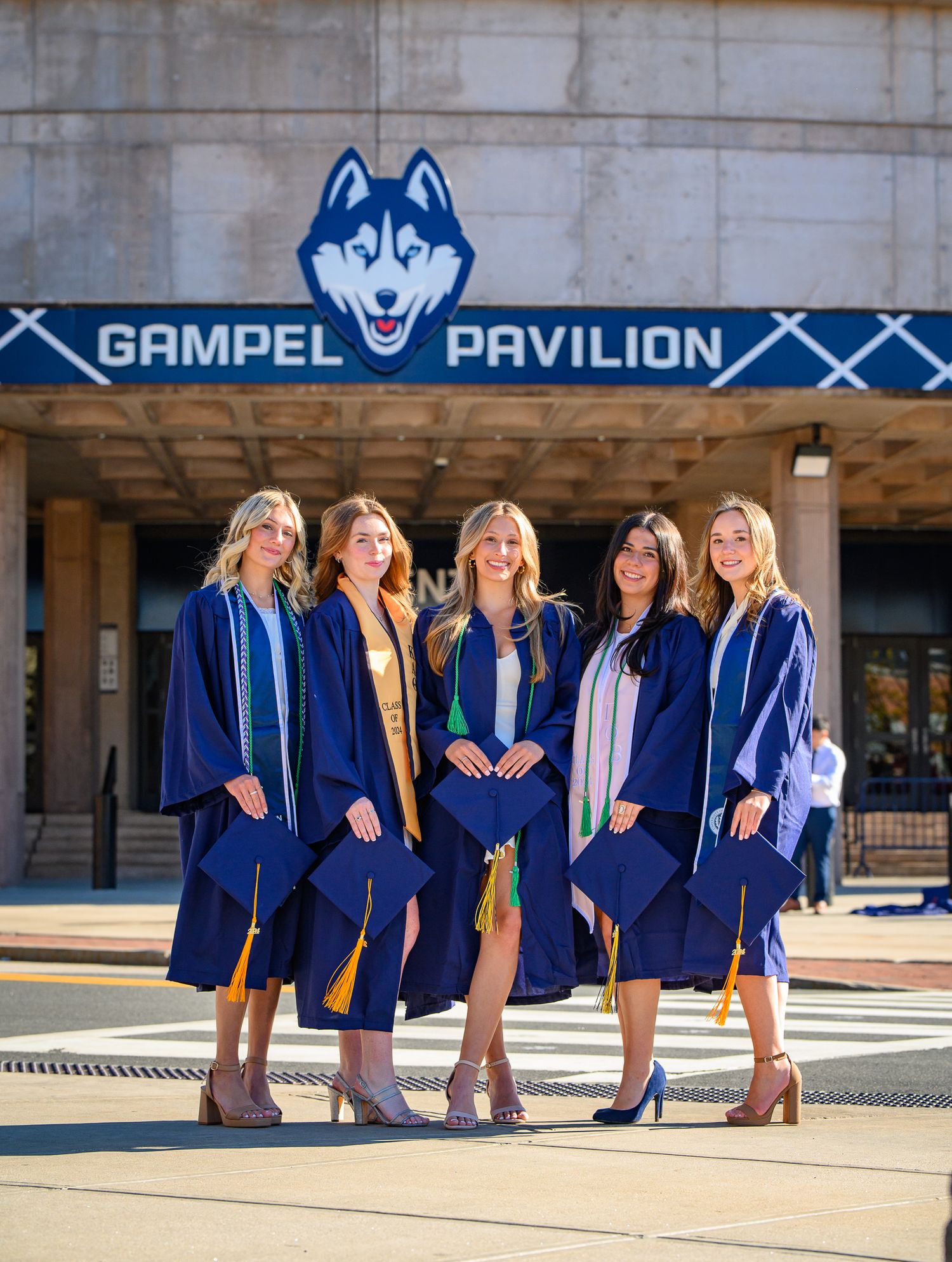 Group of graduates in blue academic regalia pose together in front of the UConn Gampel Pavilion entrance.