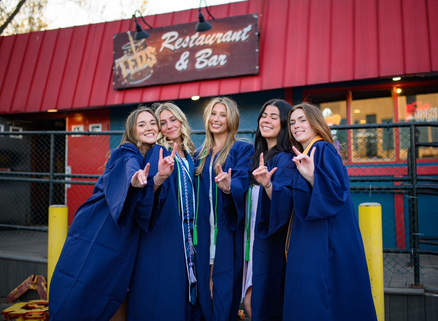 Group in blue graduation gowns make peace signs while posing outside a restaurant with red awning.