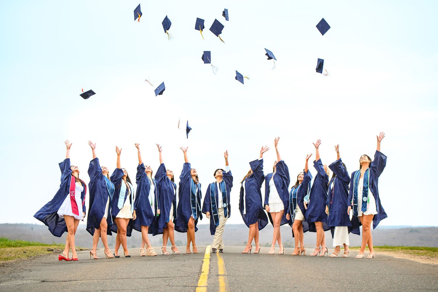 Group in matching navy blue graduation gowns toss their mortarboard caps into the air while celebrating on empty road.