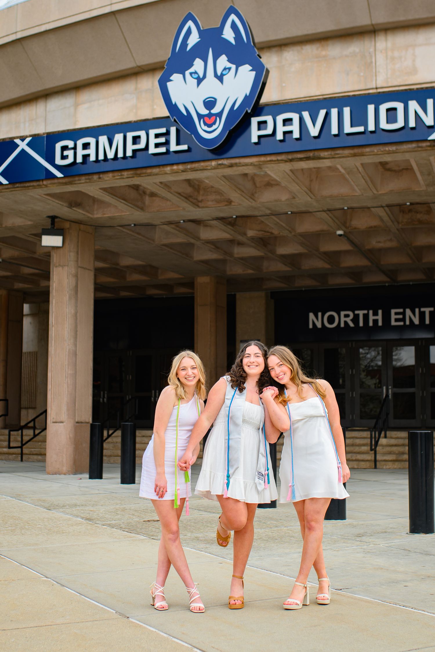 Three people in light summer dresses pose outside the Gampel Pavilion entrance with UConn Husky logo above.