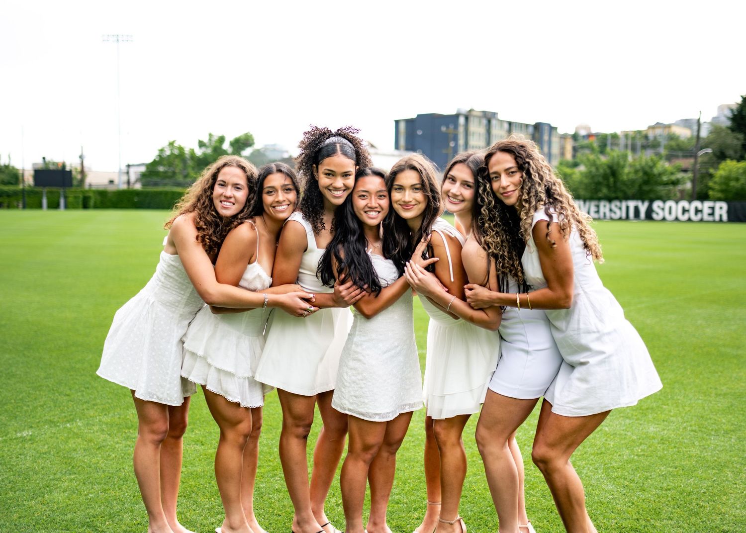 Seven young women in white dresses pose together smiling on a soccer field with city buildings in the background.