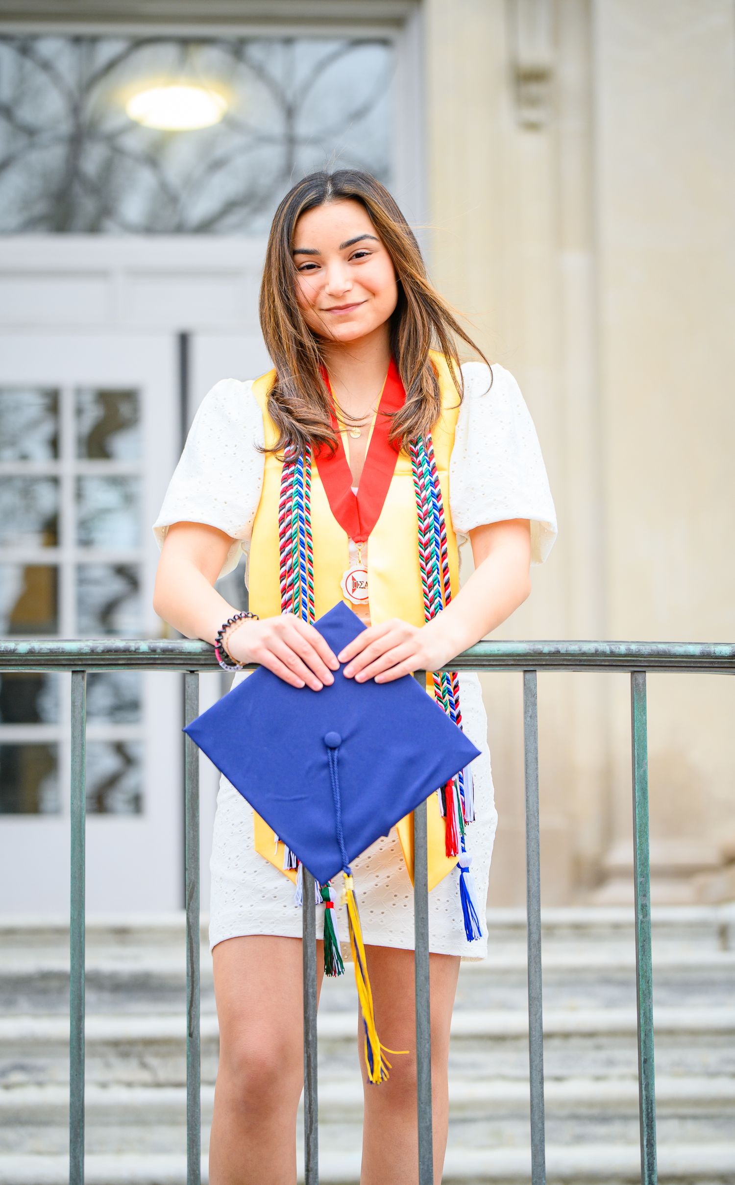 Graduate holds a blue graduation cap and wears honor cords while standing on campus steps in white dress.