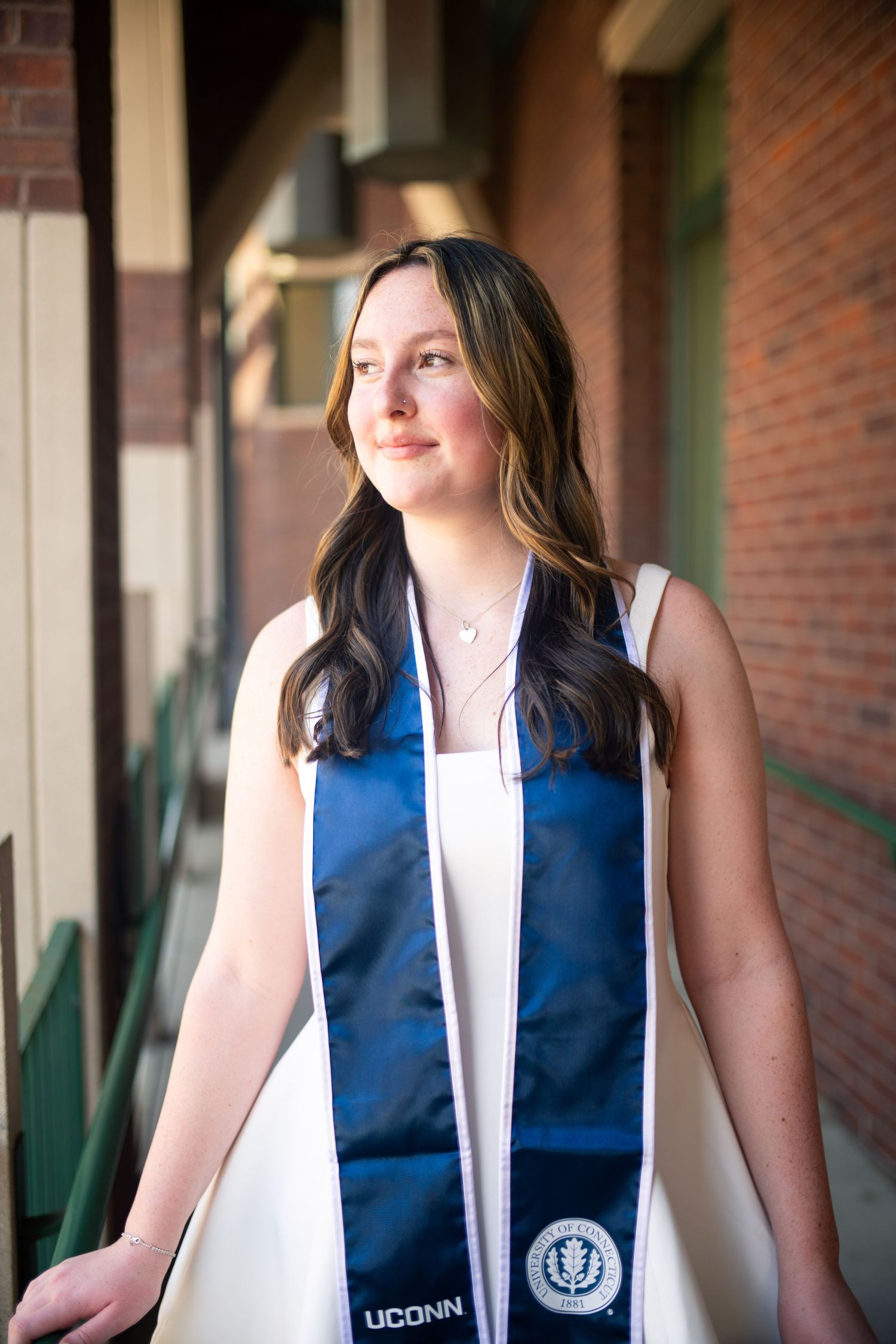 Graduate wearing blue and white UConn graduation stole standing outside brick building in natural lighting.