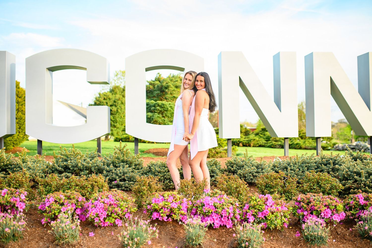Two people pose in front of large PENN letters surrounded by colorful flower gardens on a sunny day.