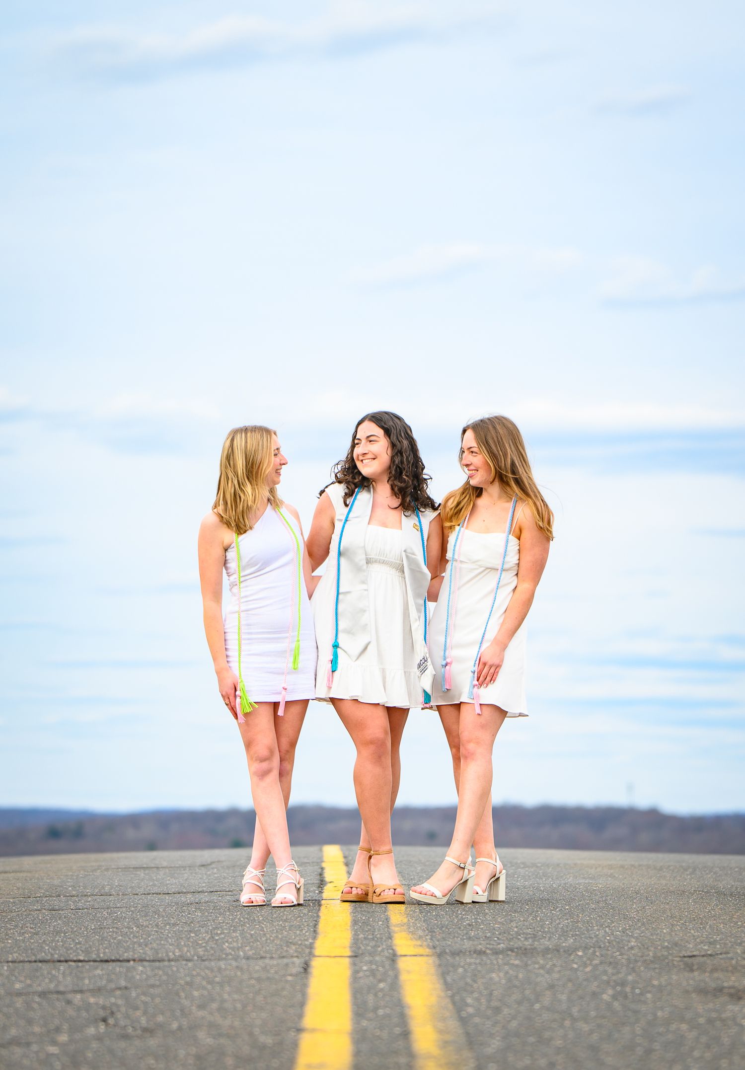 Three women in white dresses stand on a yellow line painted on an empty road under a blue sky.