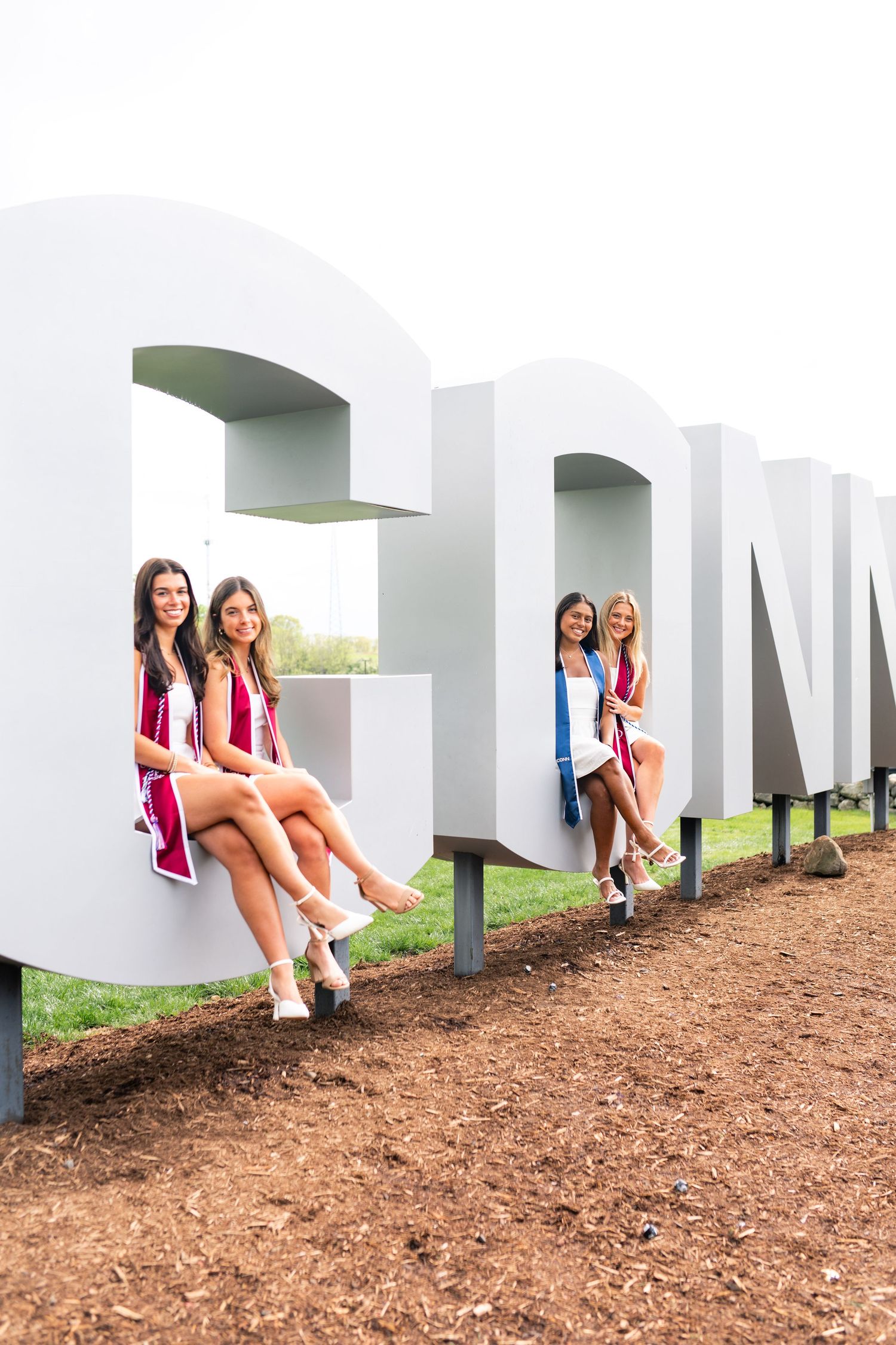 Students sit and pose on large white letters spelling out a word on a hill with scattered mulch on the ground.