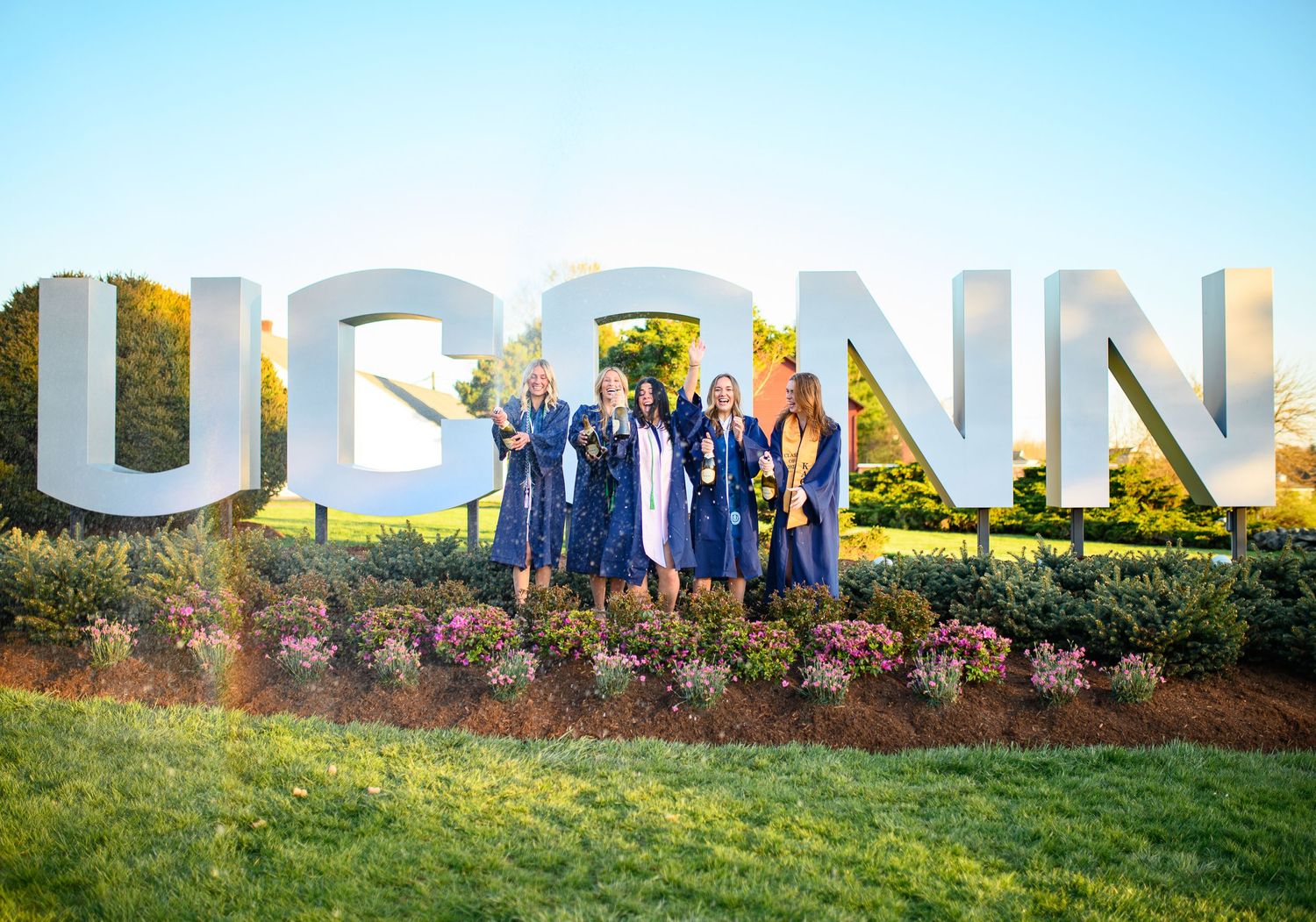 A group in graduation gowns poses in front of large white UCONN letters on a grassy hill.