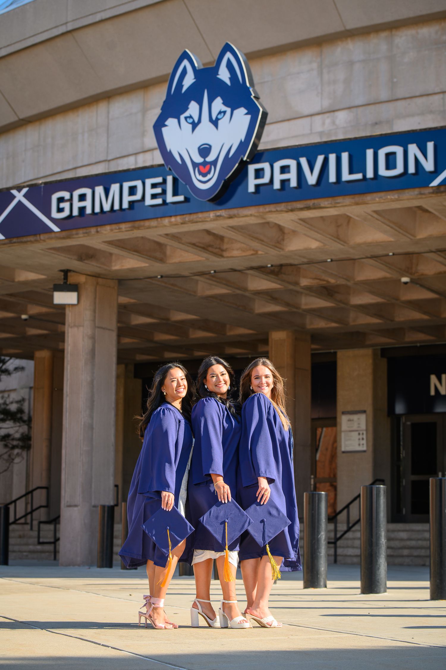 Three graduates in blue commencement robes pose together outside Gampel Pavilion with UConn Husky logo visible above.