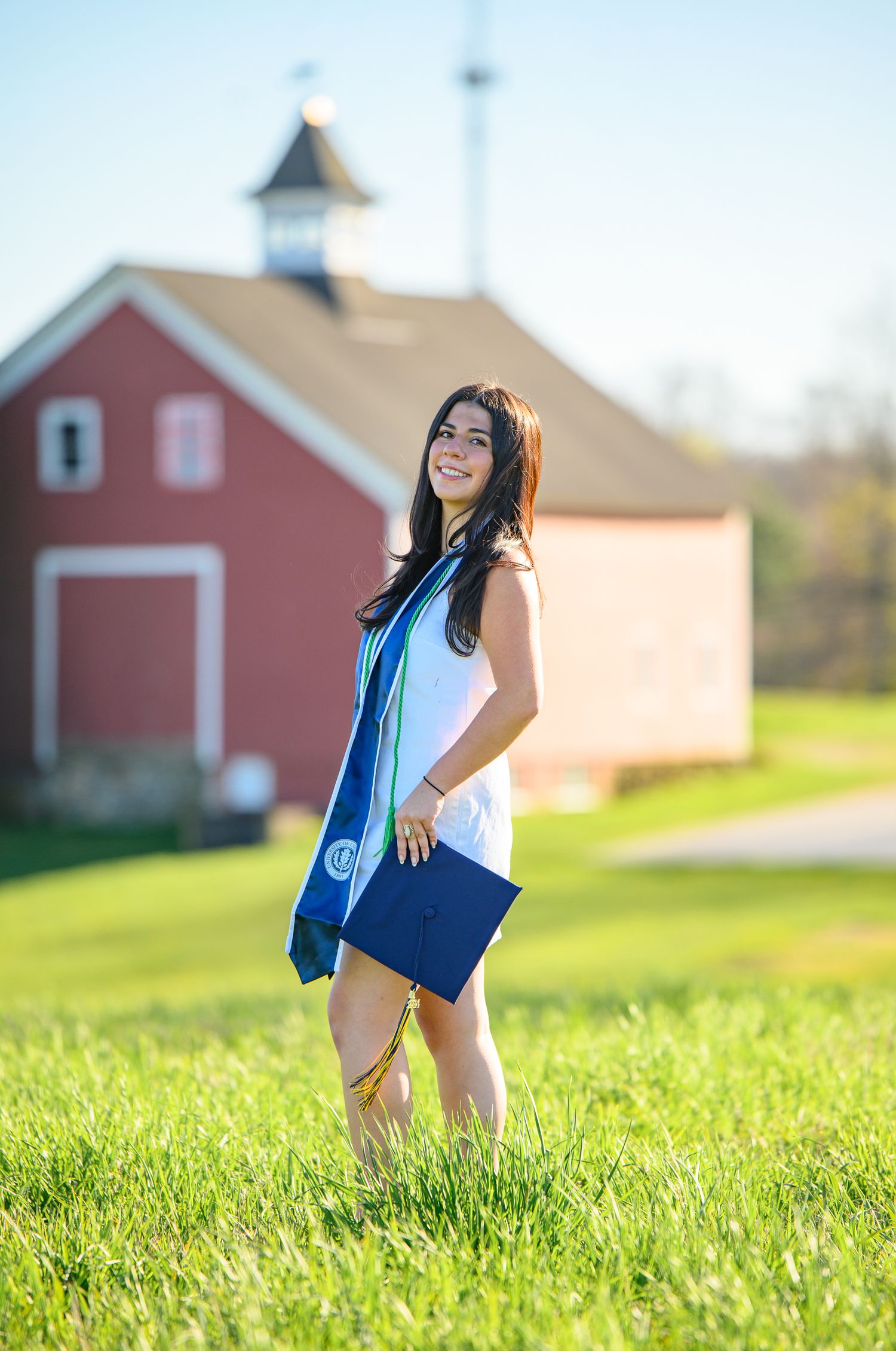 Graduate in white dress and blue sash stands in a grassy field with a red barn in the background.