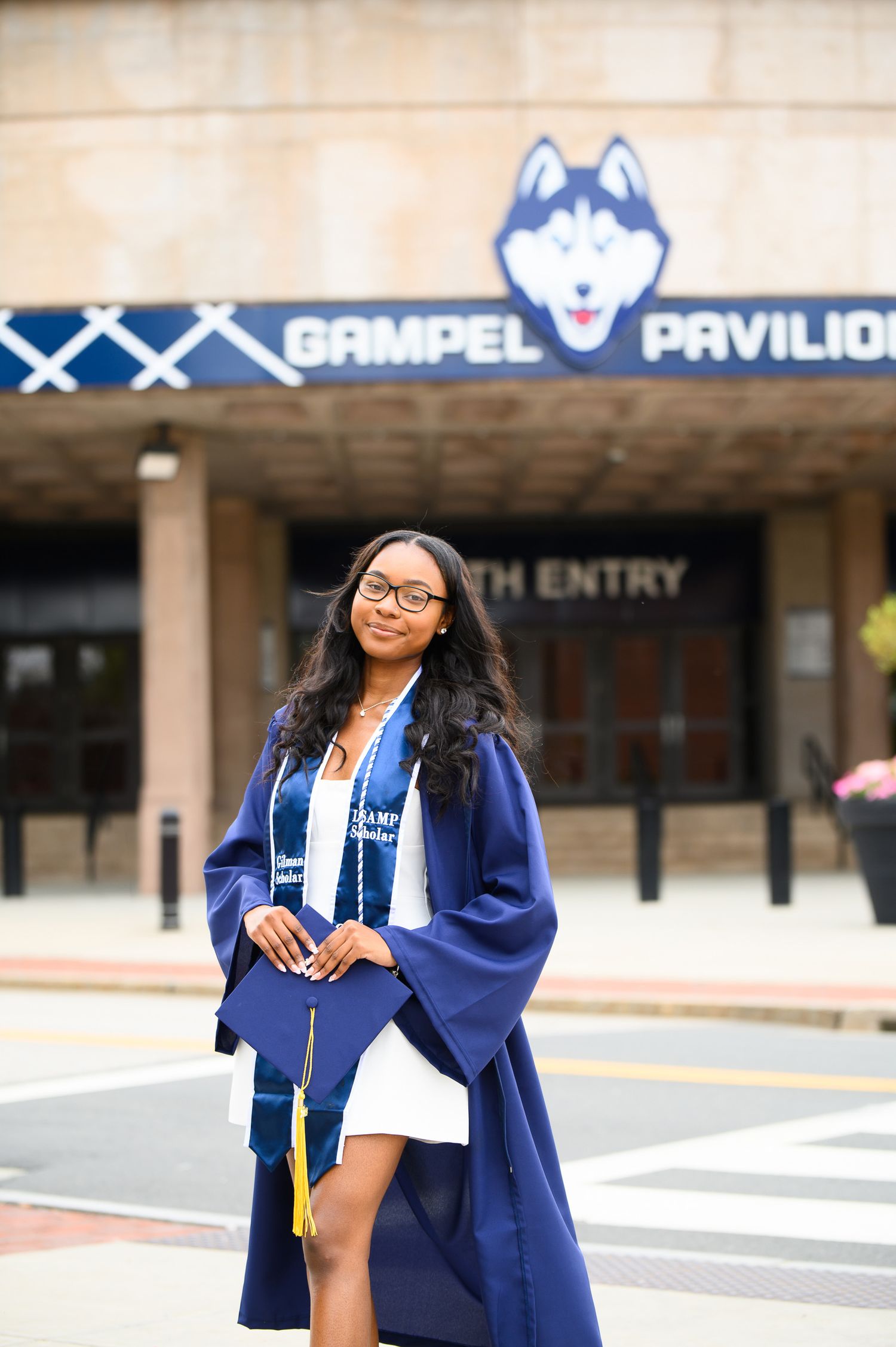 Graduate poses in blue cap and gown outside Gampel Pavilion at UConn with the husky mascot logo visible.