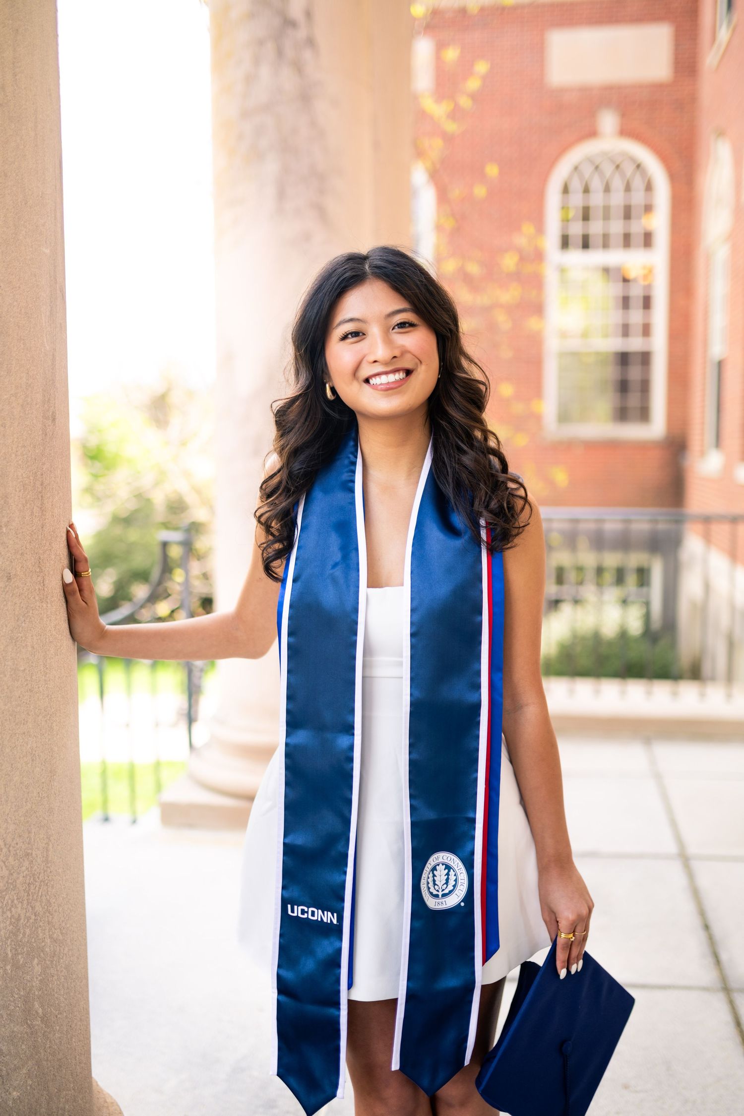 Graduate wearing blue and white graduation stole and holding diploma stands smiling by brick building with arched window.