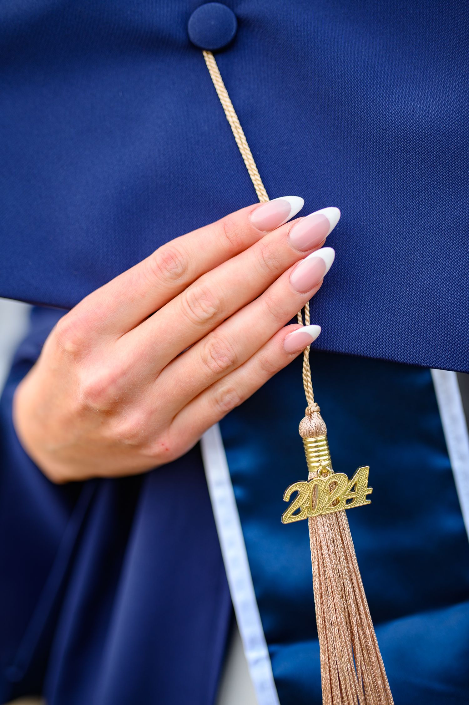 Close-up of a hand with white manicured nails touching a gold 2024 graduation tassel against a navy blue background.