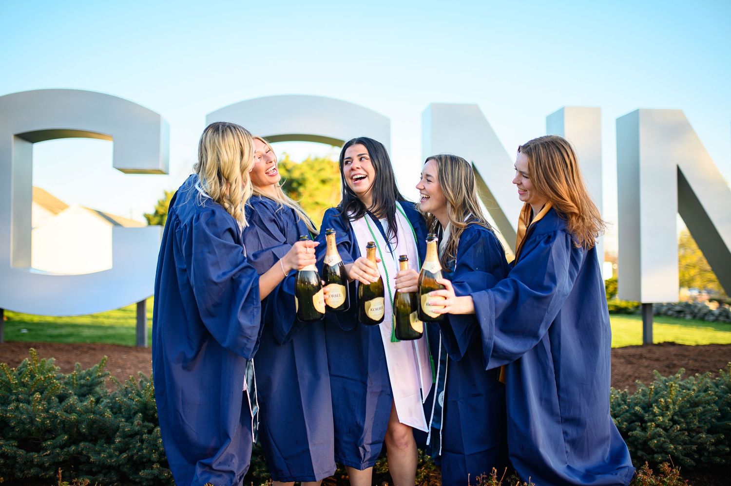 A group of graduates in navy blue robes celebrate with champagne bottles in front of large outdoor letters.