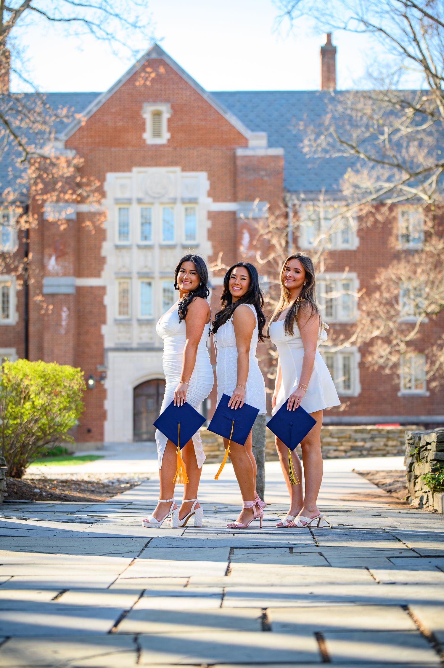 Three graduates in white dresses pose with blue diplomas on campus steps in front of brick university building.