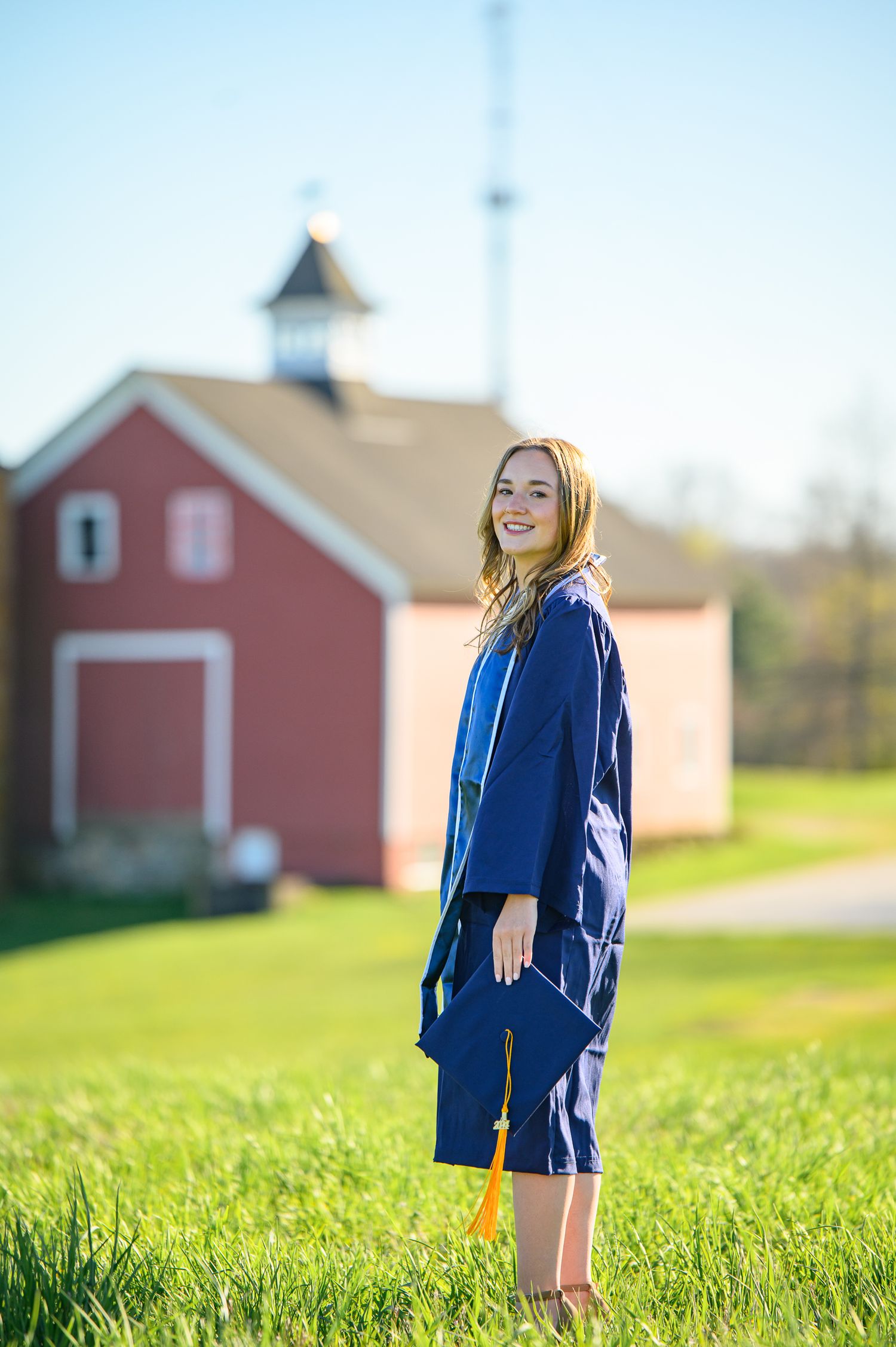 Graduate in blue cap and gown smiles while standing on grassy hill with red barn in background on sunny day.