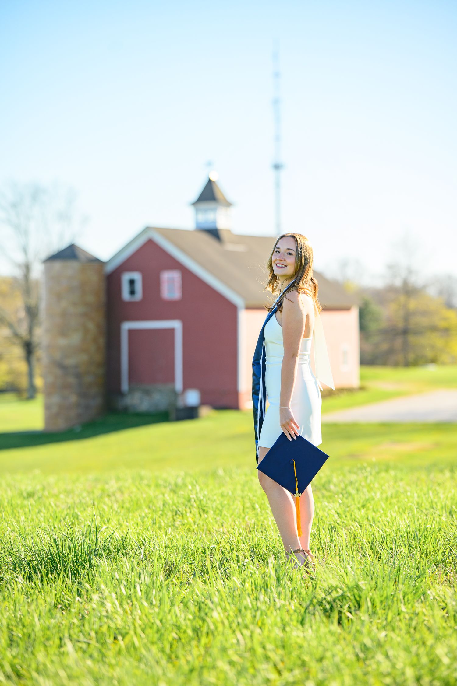 A sunny day with a barn and silo in the background and a graduate posing in a white top on green grass.