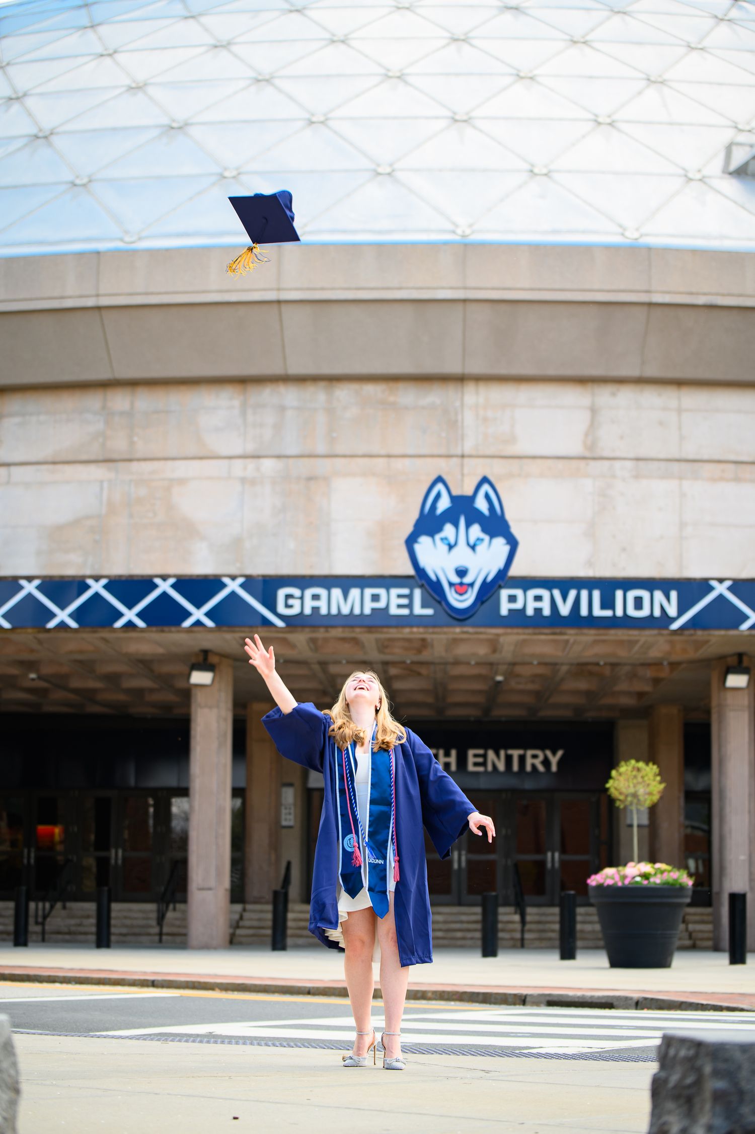 Graduate celebrates in cap and gown outside the Gampel Pavilion basketball arena at UConn.