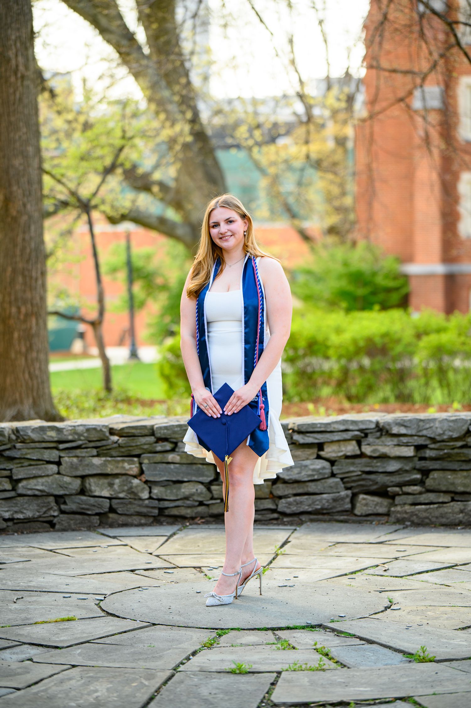 Young woman in white dress and blue cardigan poses for photo on stone wall in springtime garden setting.