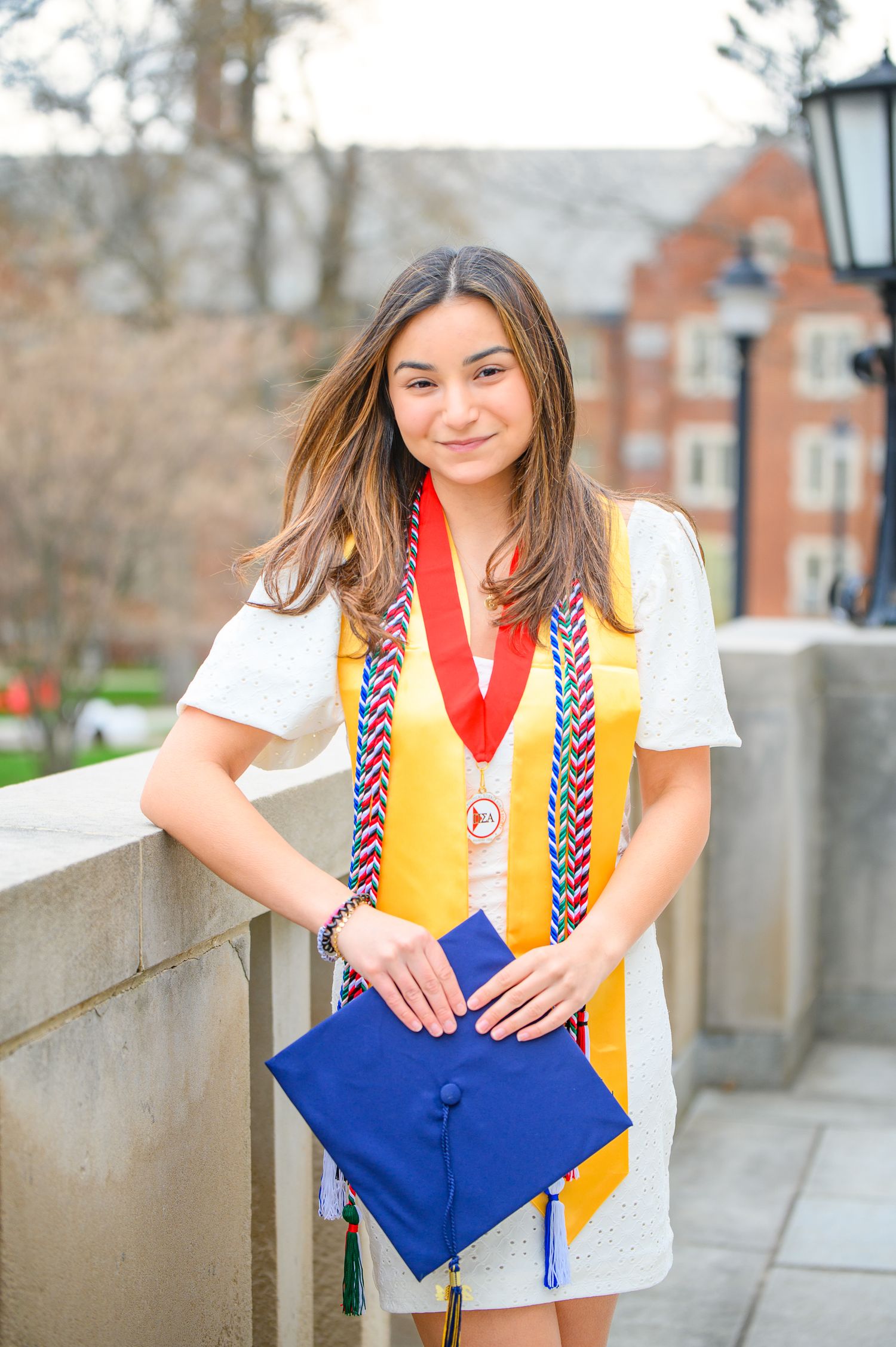 Graduate in white dress and gold stole holds blue graduation cap while posing on campus balcony.