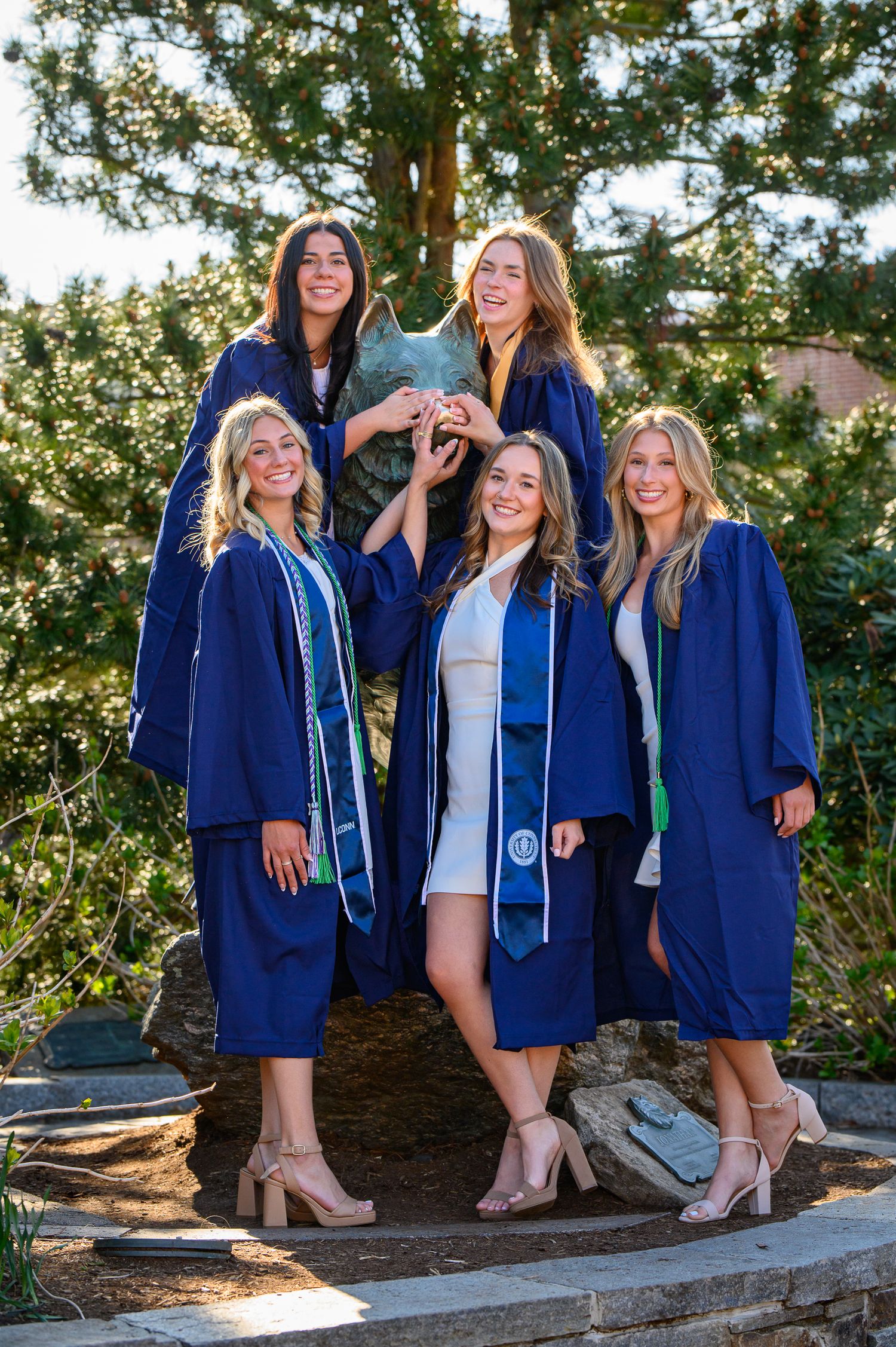 A group of graduates in navy blue caps and gowns pose together outdoors on a sunny day among trees.