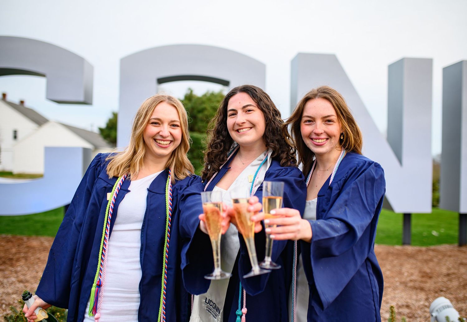 Three graduates in blue gowns celebrate with champagne glasses in front of large outdoor letters.