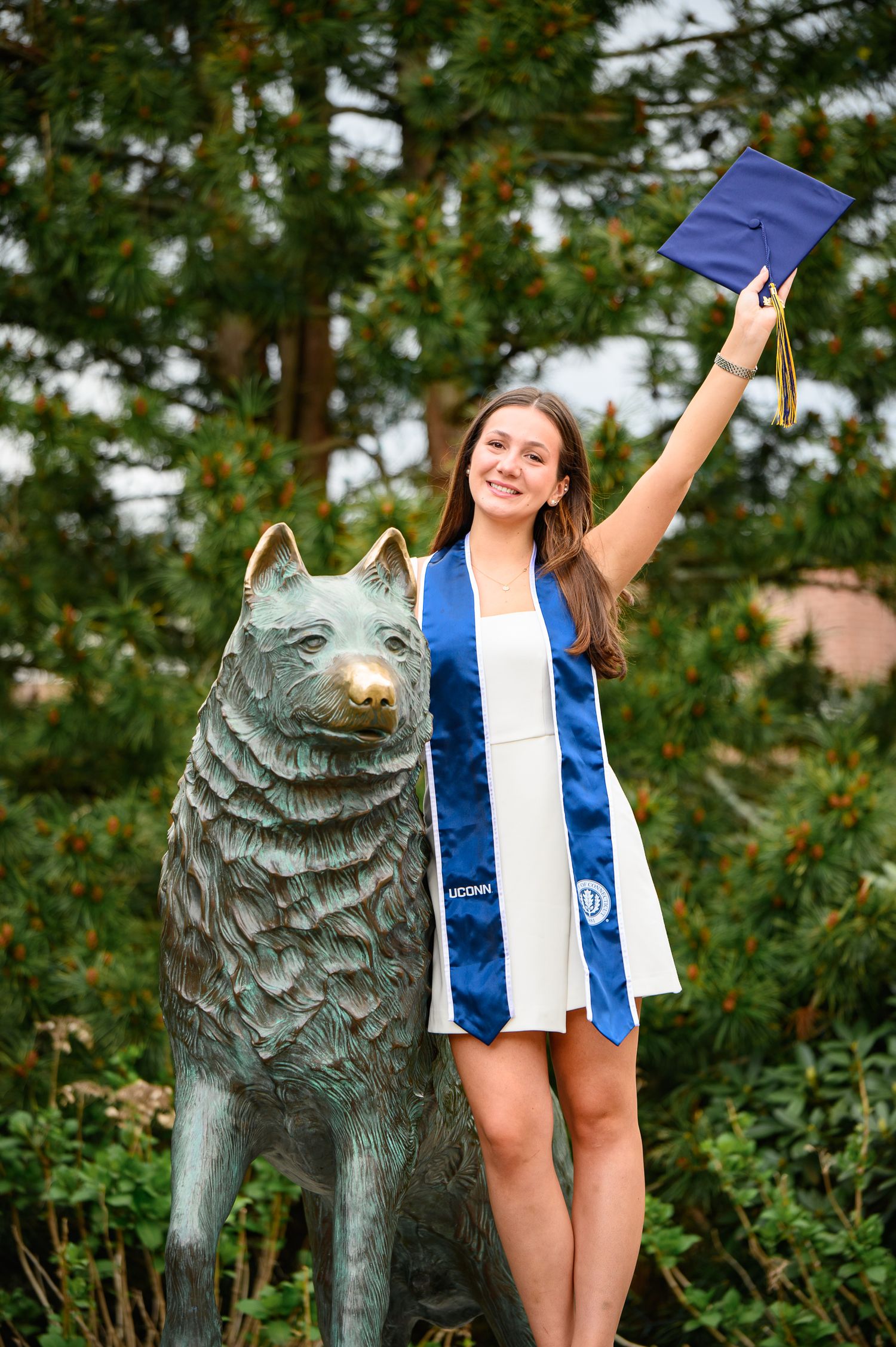 A graduate in blue and white regalia celebrates next to a wolf statue on campus while holding up their cap.