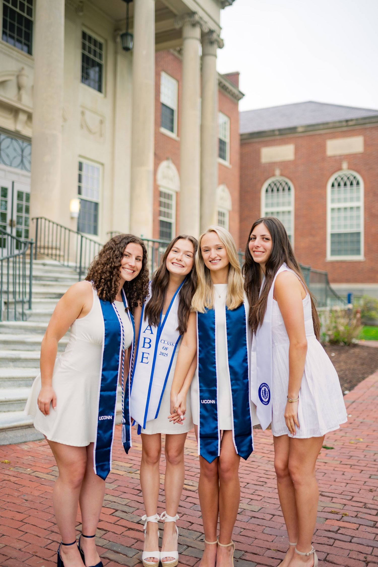 Group in blue graduation stoles pose together for celebration photos on brick pavement outside a collegiate building.
