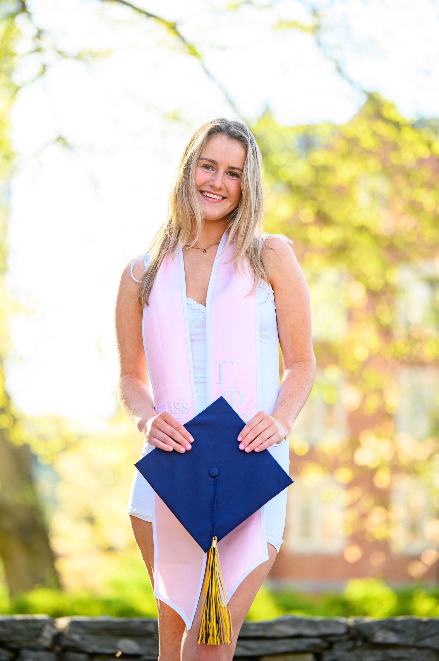 Graduation photo shows someone in a white outfit holding a blue cap with gold tassel outdoors on a sunny day.