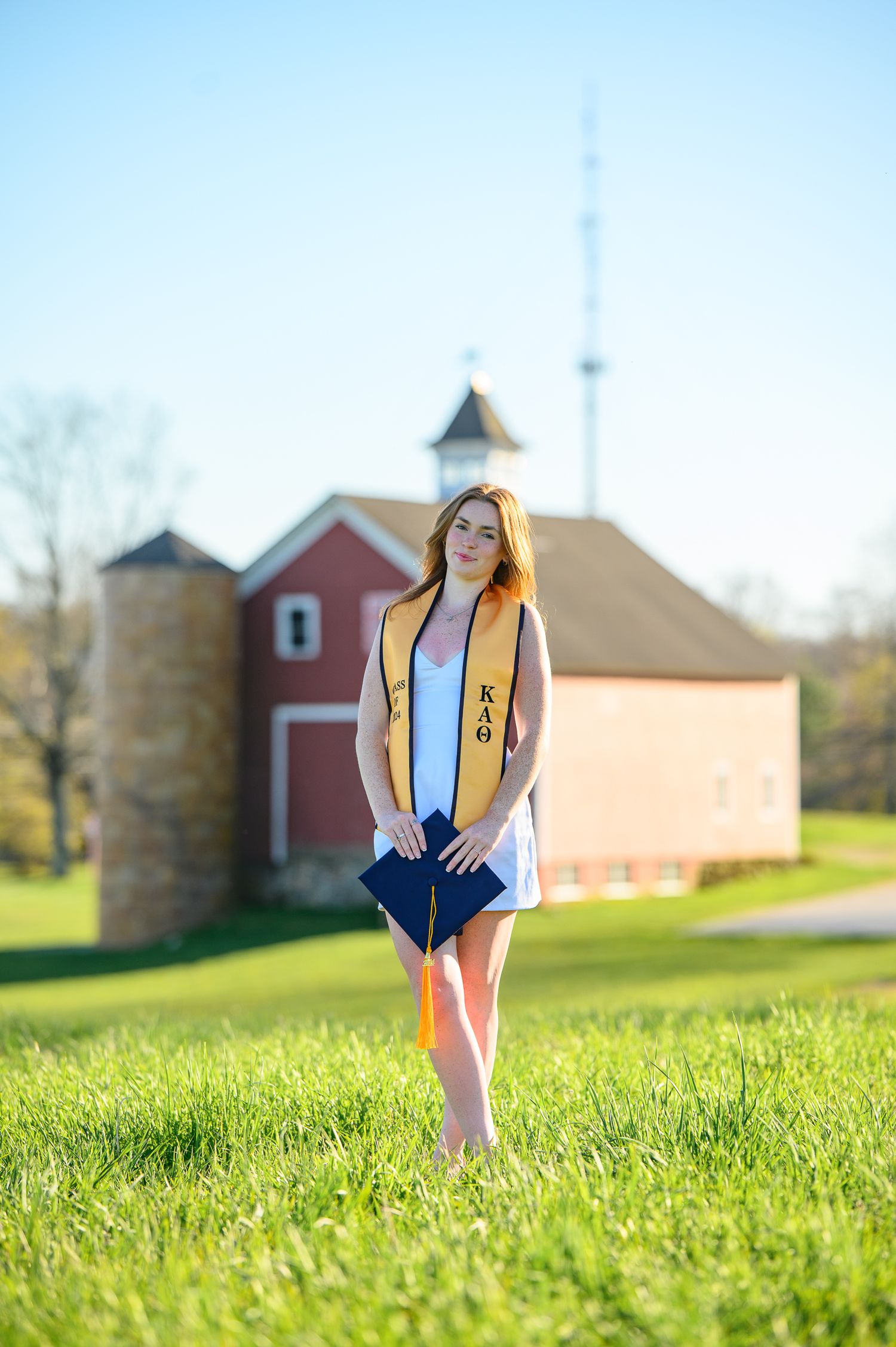 Graduate in gold honors stole stands on sunny grass field with red barn and silo in background.