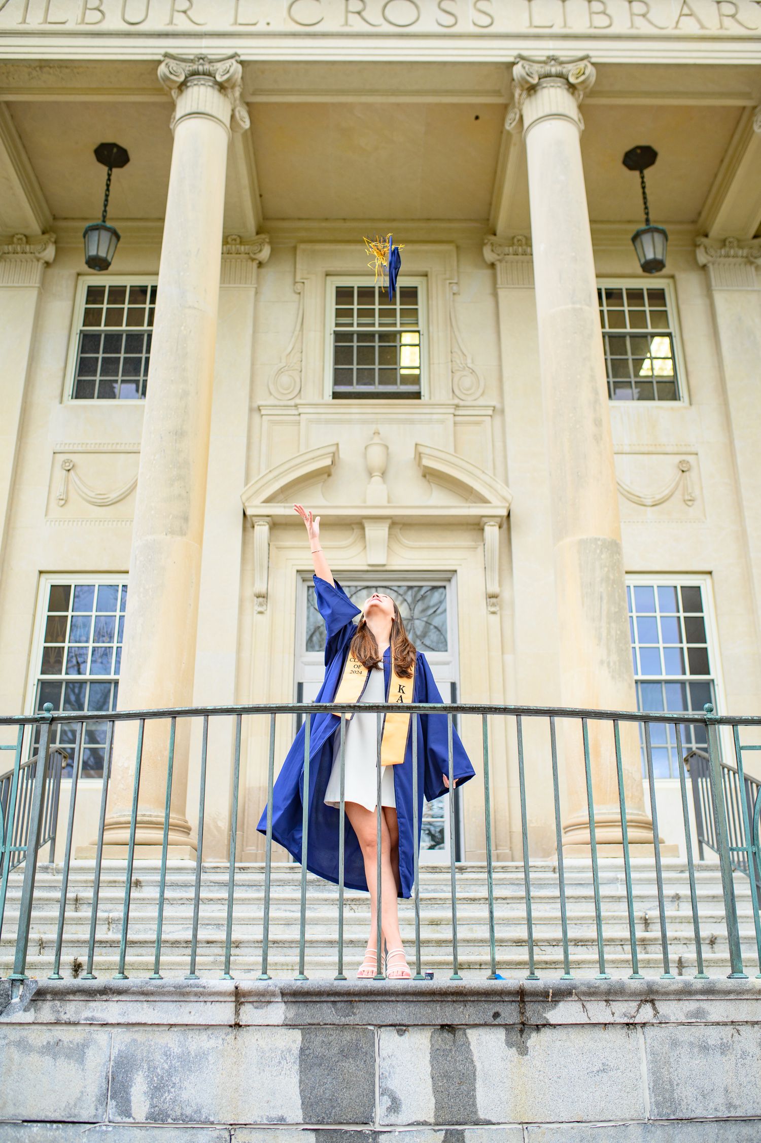 A graduate in blue cap and gown poses on the steps of the Wilbur L. Cross Library with classical columns and architecture.