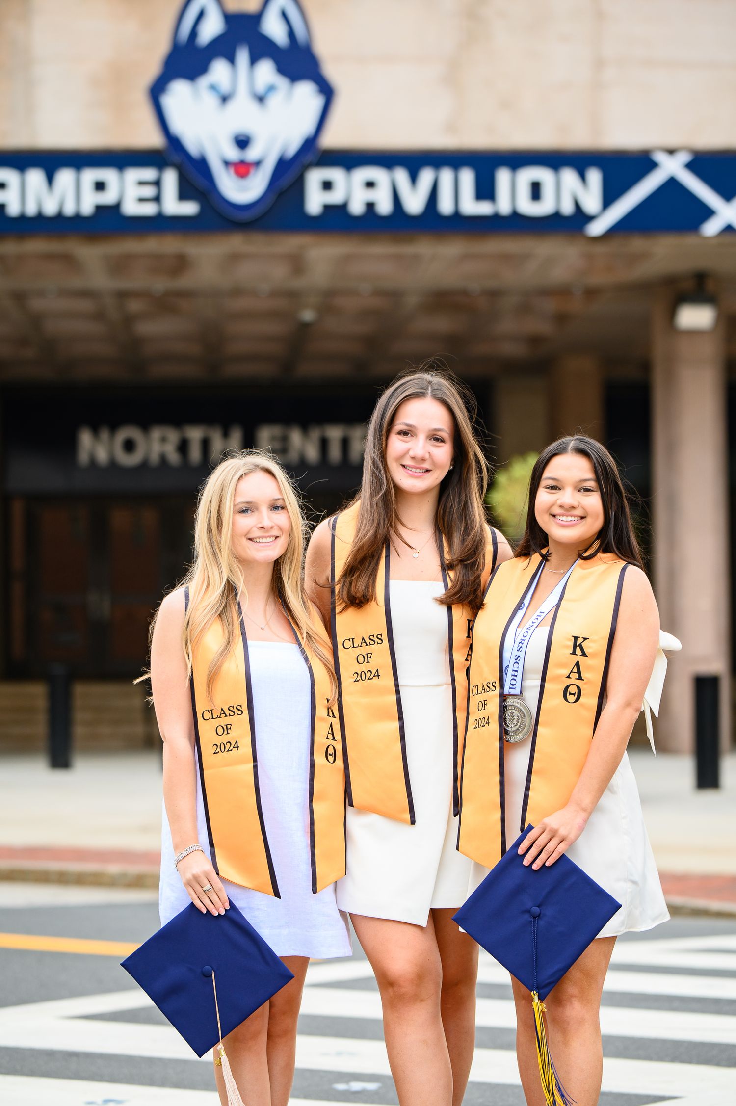 Three graduates in white dresses and gold KAO stoles hold blue diplomas in front of Gampel Pavilion at UConn.
