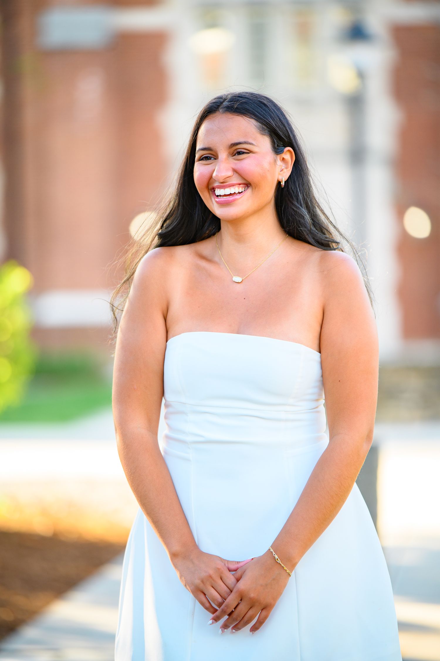 Smiling person with long dark hair wearing a white strapless dress stands outdoors on a sunny day.