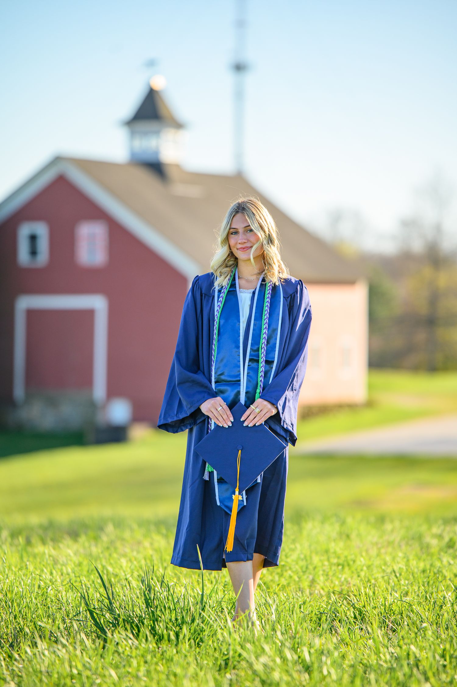 Graduate in royal blue cap and gown stands in grassy field with red barn in background during sunny day.