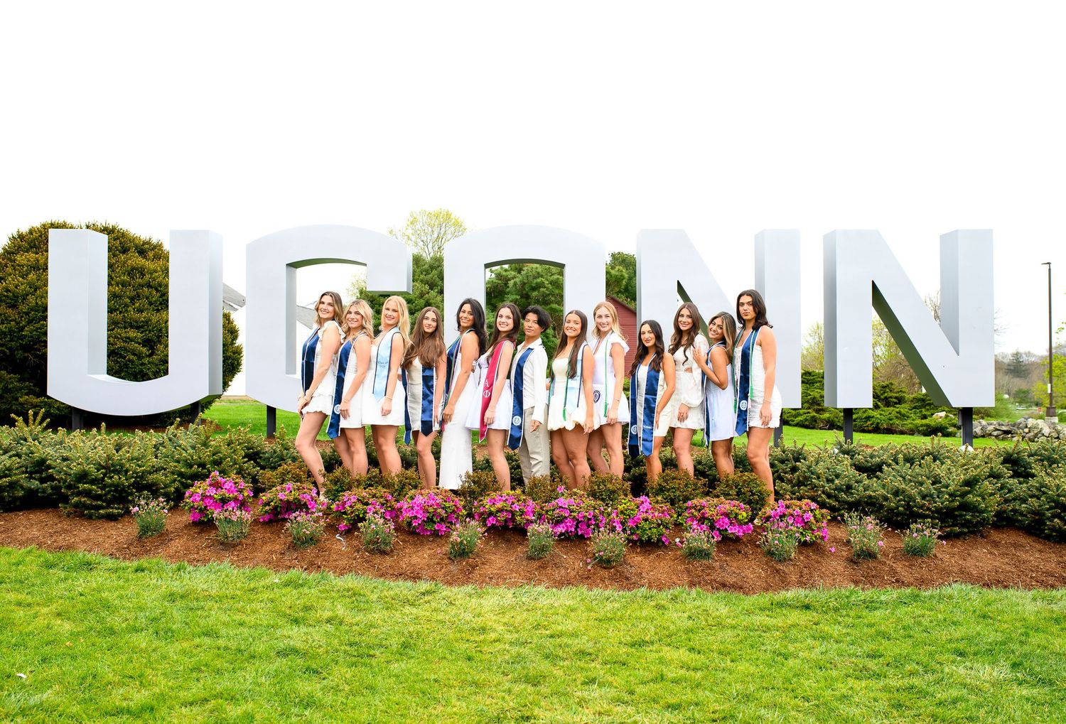 Group of cheerleaders pose in white uniforms in front of large white UCONN letters and flower garden.