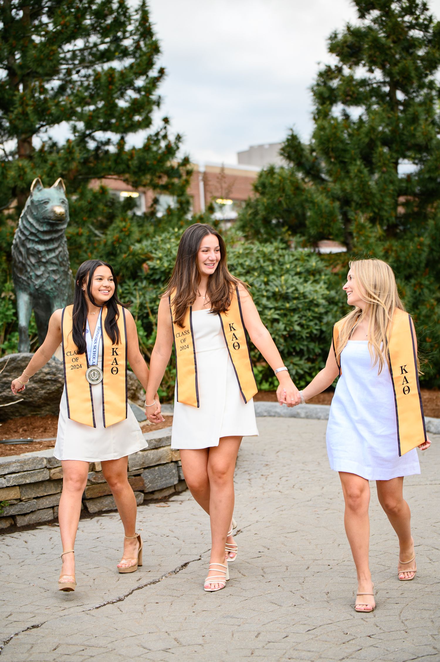 Three students wearing gold graduation stoles and white dresses walk together on a path with trees and buildings in background.