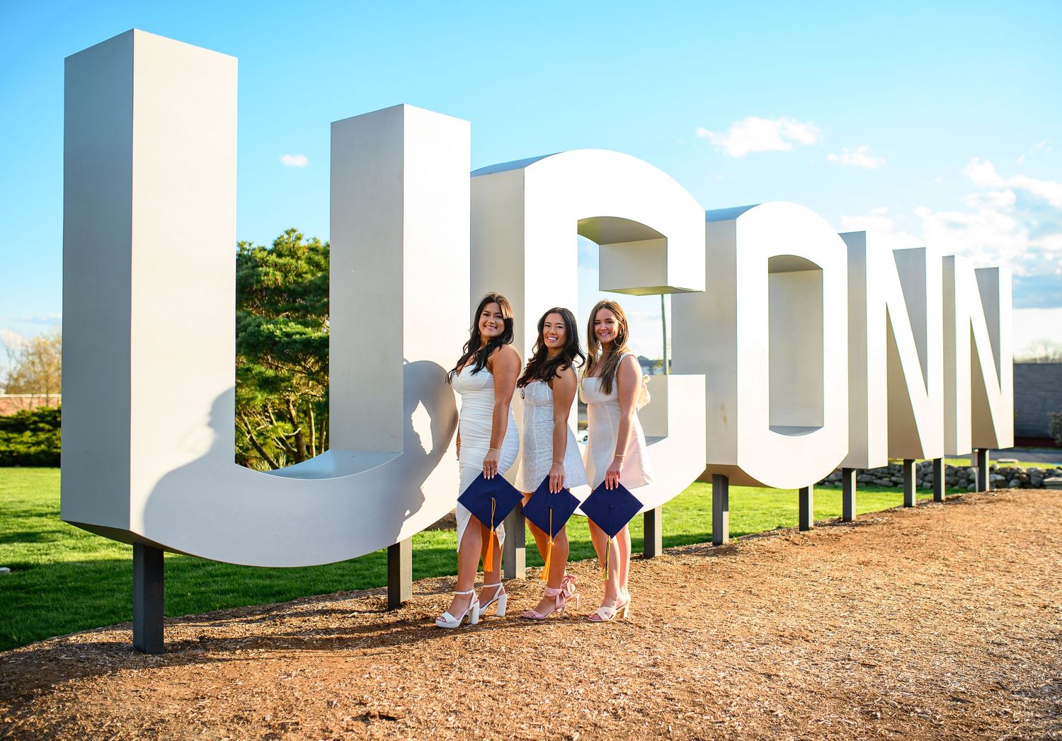 Three people pose together in front of large white UCONN letters on a sunny day.
