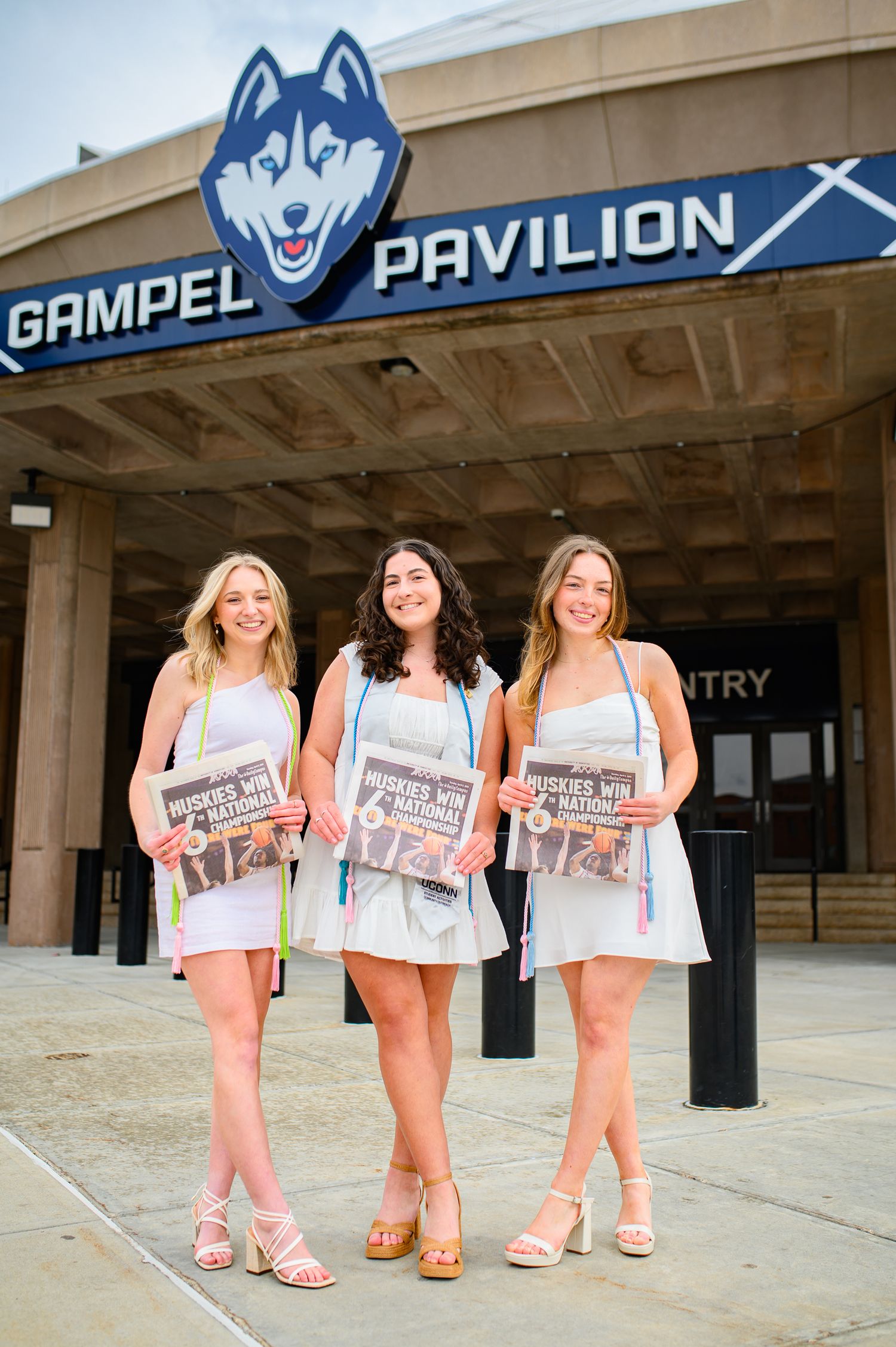 Three students in light summer dresses hold newspapers while standing in front of the UConn Gampel Pavilion entrance.