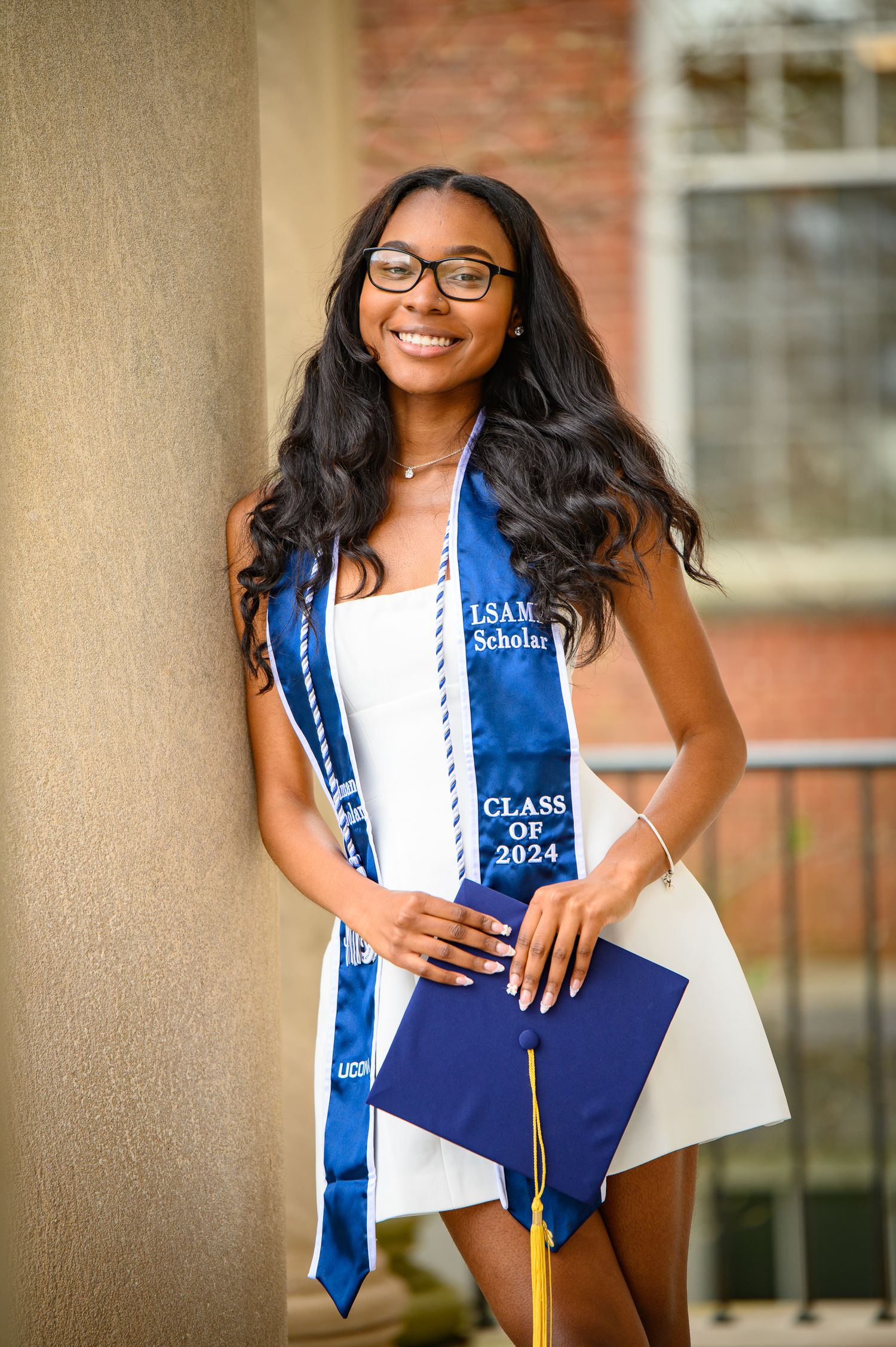 Graduation photo showing smiling graduate in blue and white sash with Class of 2024 stole holding diploma case against brick wall.