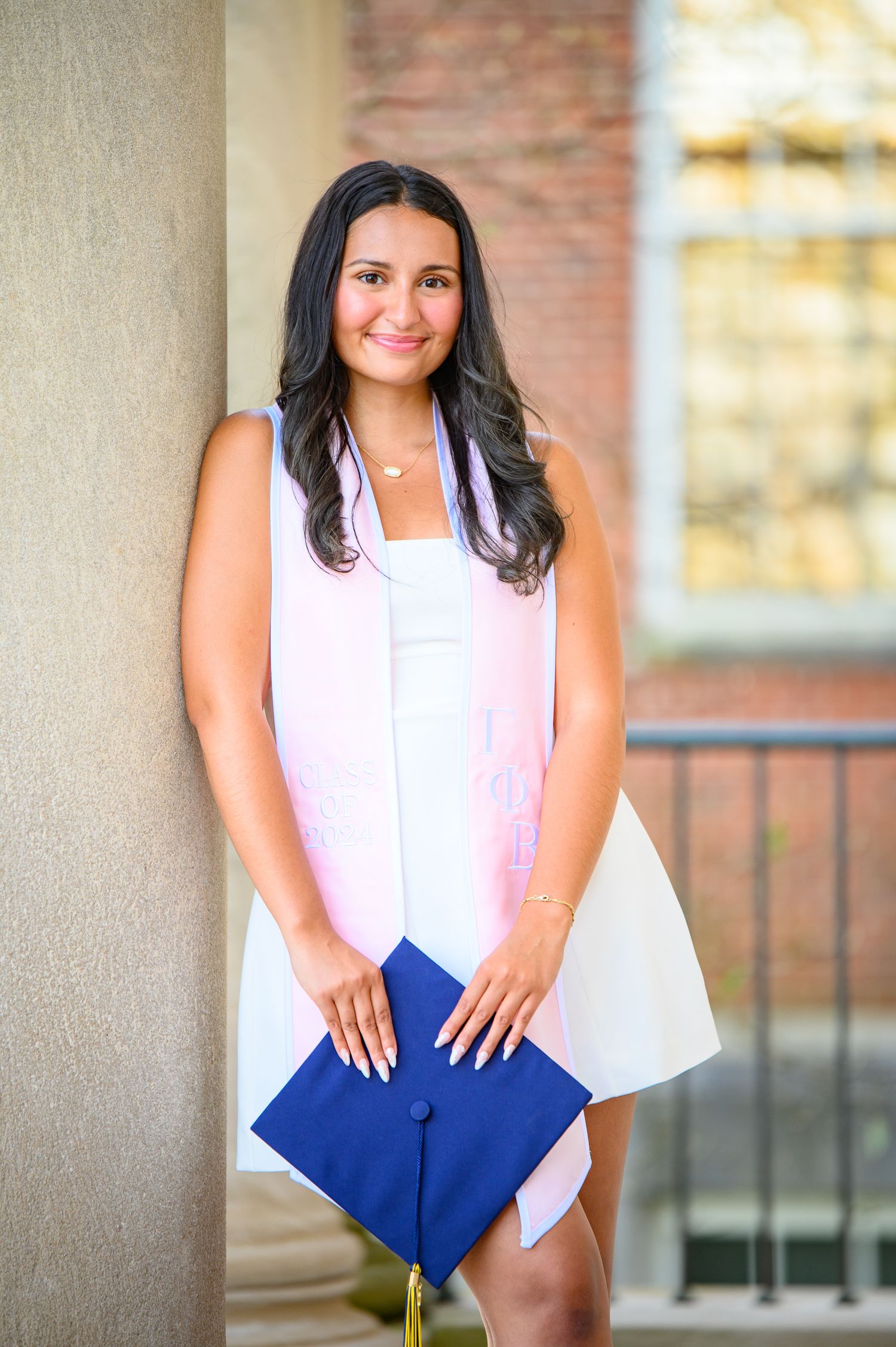 Graduate in white dress holds blue diploma cover while standing against brick building exterior during photo session.