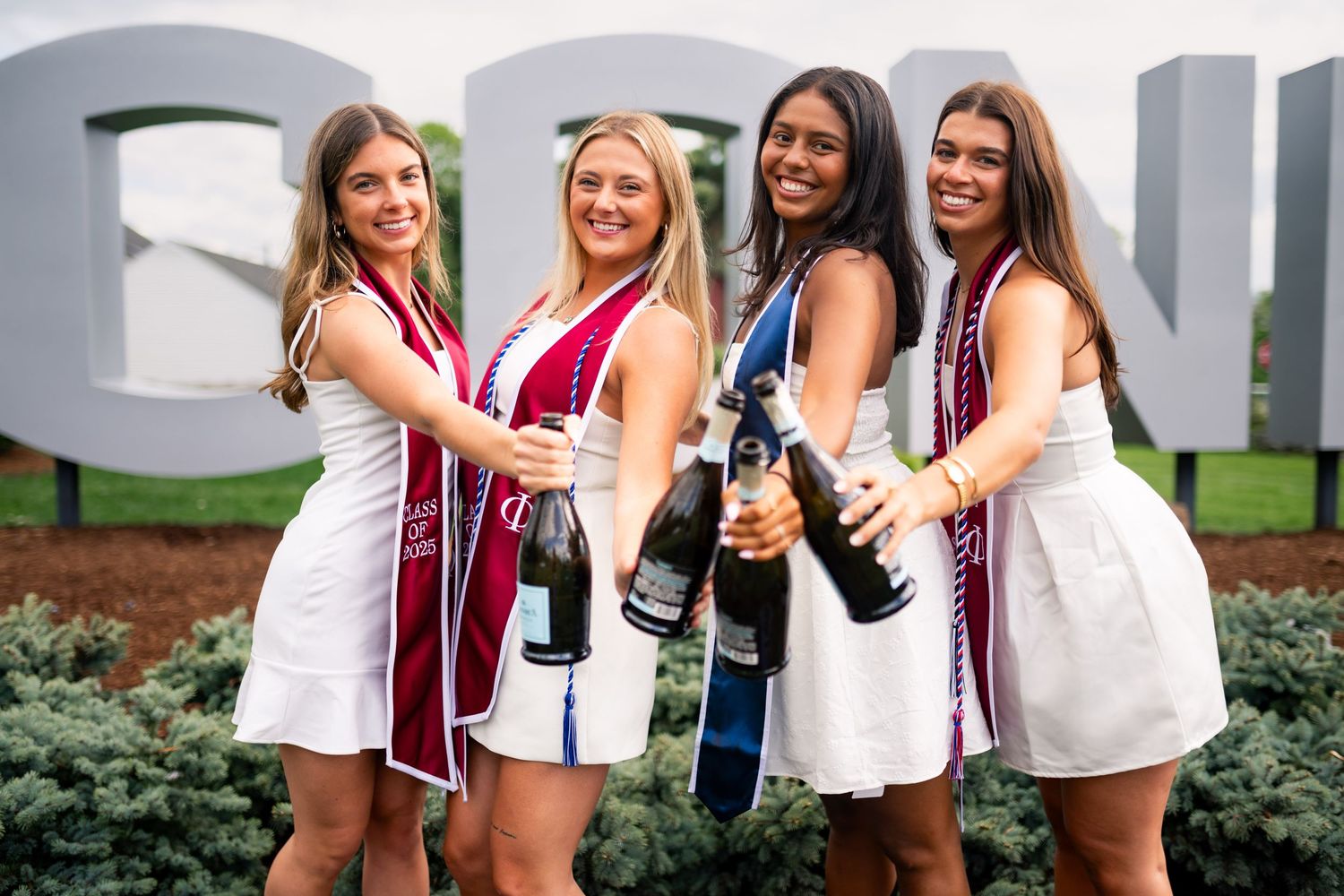 A group of cheerleaders in white uniforms stand together smiling and posing for a photo outdoors.