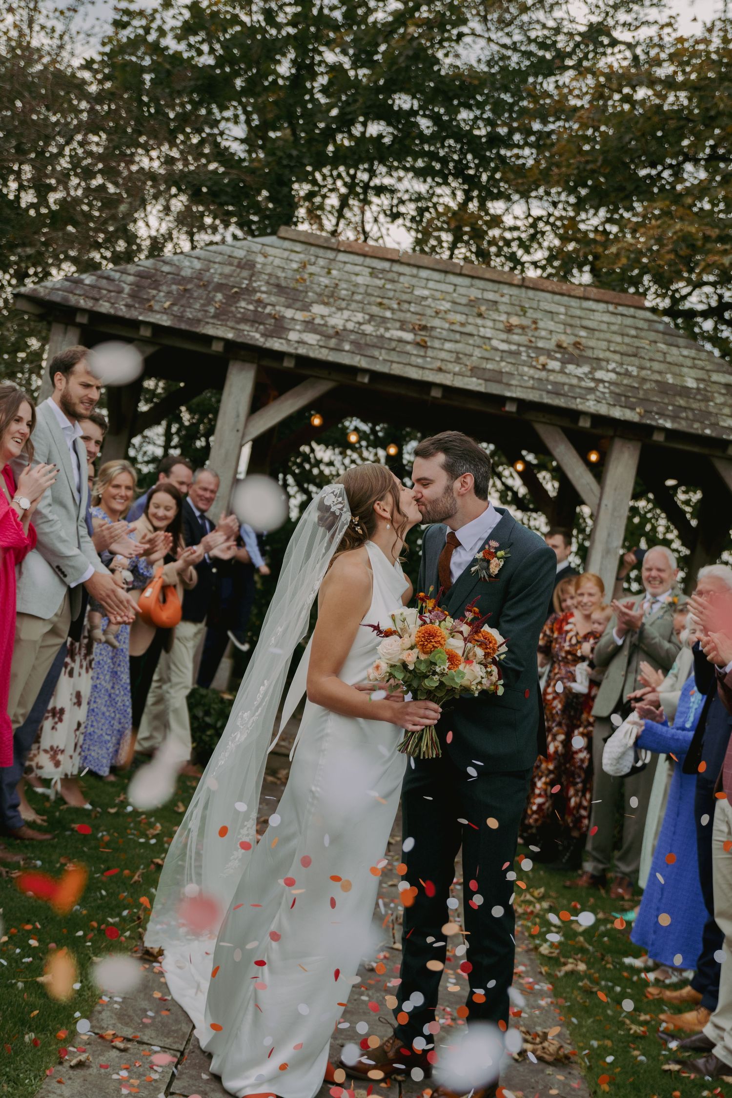Wedding guests shower newlyweds with colorful autumn leaves and flower petals under a rustic wooden gazebo.