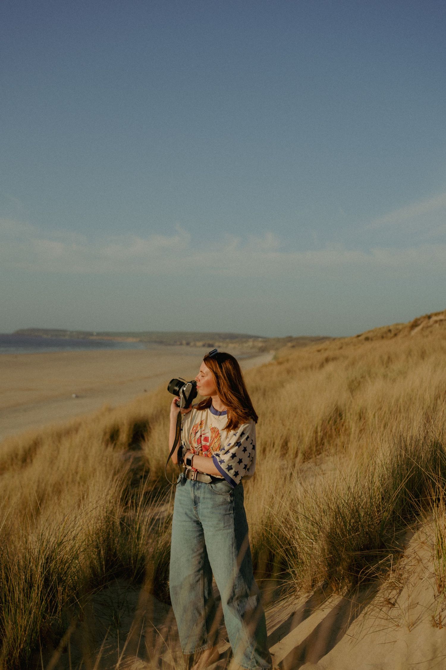 Woman photographer standing on sandy beach dunes with camera, wearing jeans and patterned sweater, golden hour light.
