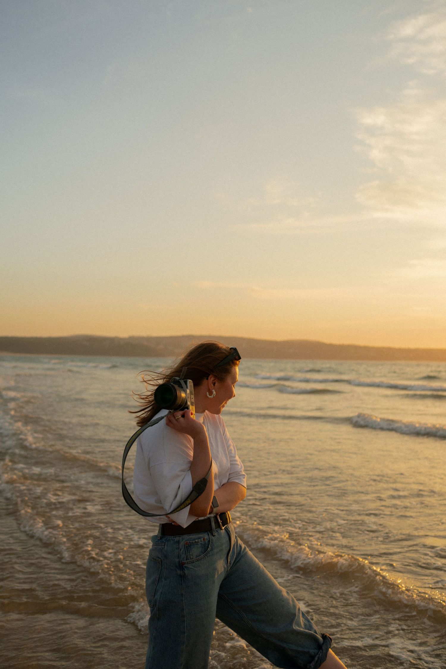 Woman in white tee and jeans walks along ocean shore at golden sunset, hair blowing in breeze.